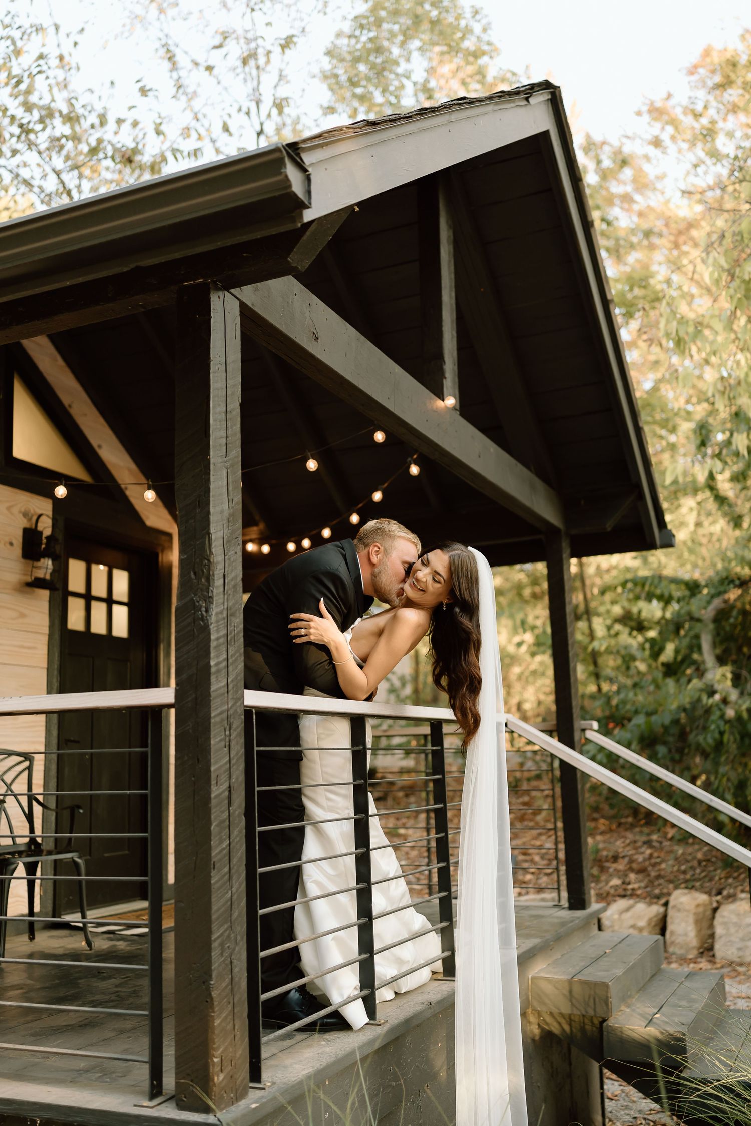 A newlywed couple shares an intimate kiss on a rustic cabin porch with string lights overhead.