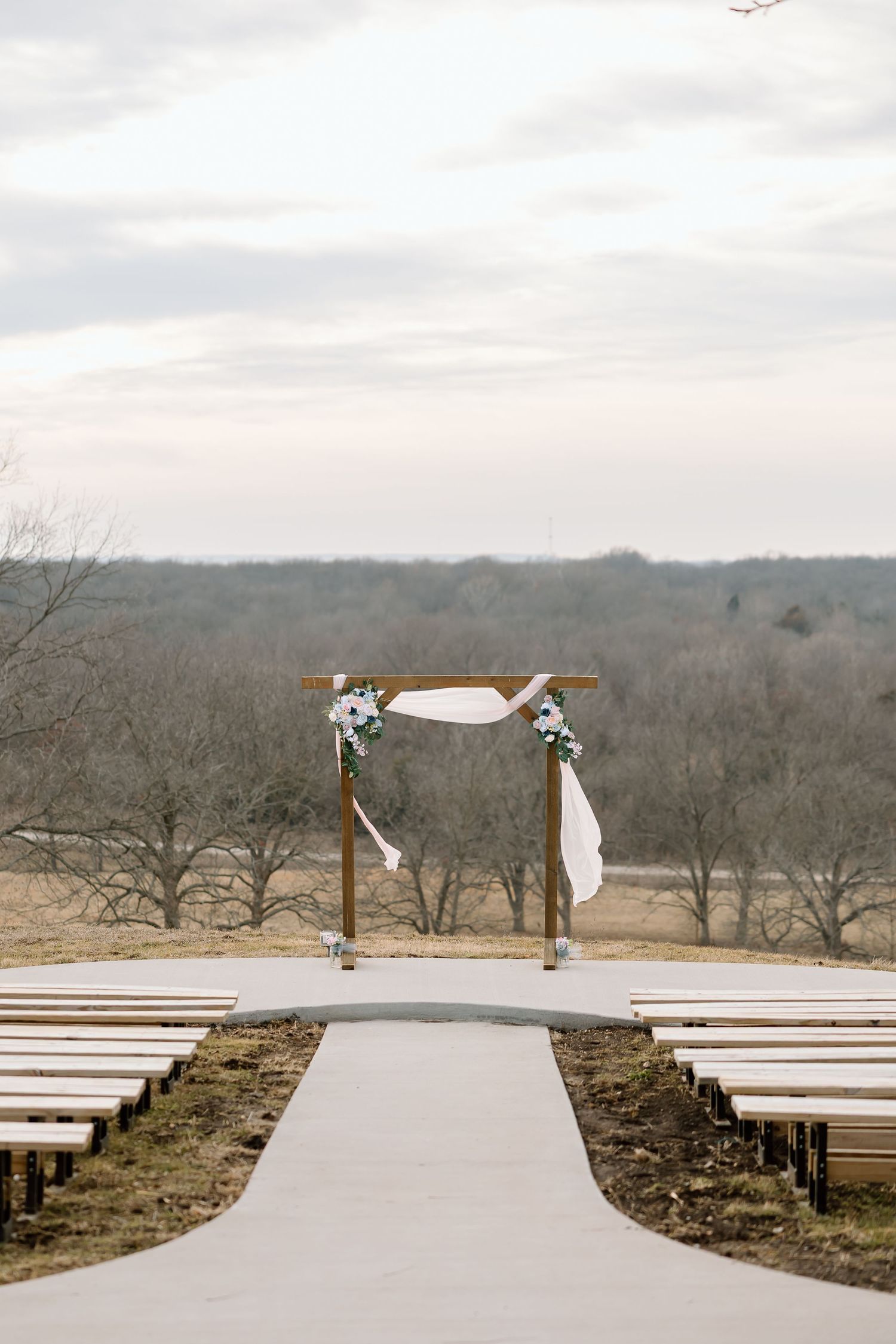 Rustic wooden wedding arch with white fabric and floral decorations stands before rows of benches on a hillside outdoor venue.