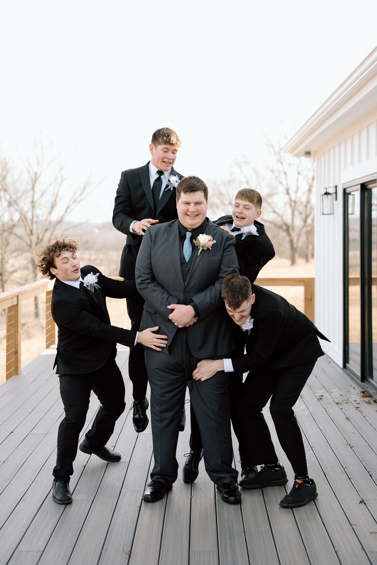 Groomsmen in black suits playfully posing in a sequence of falling down poses on a wooden deck.