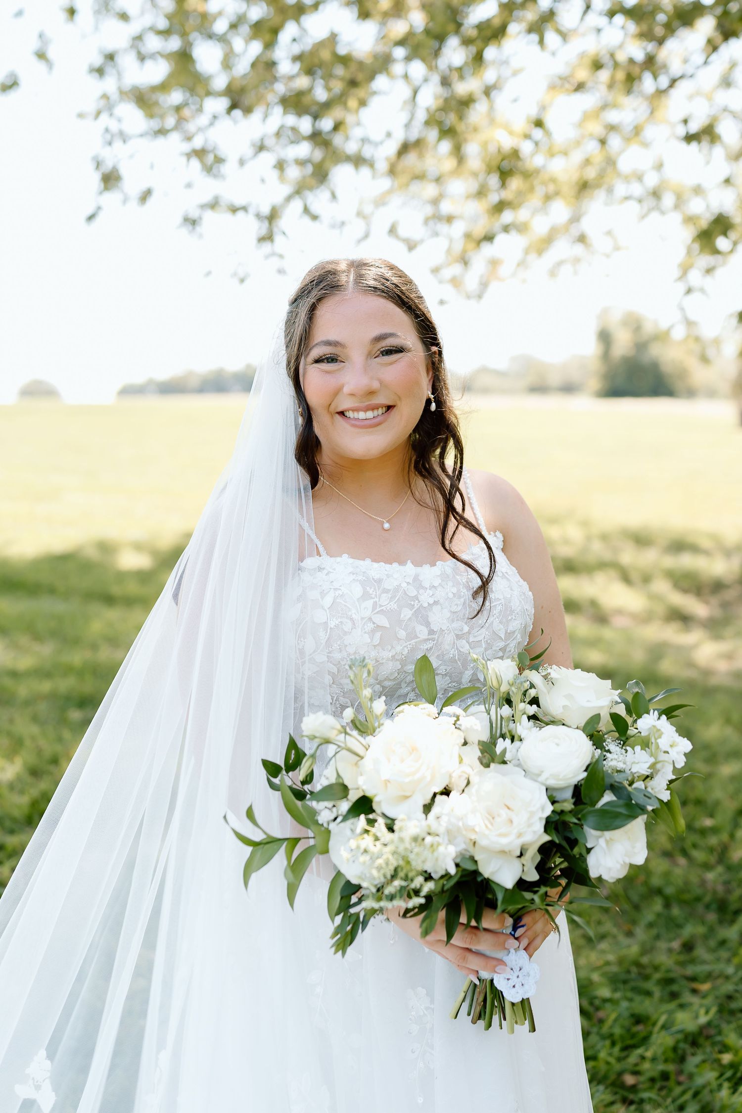 A stunning bridal portrait showcasing an elegant white wedding bouquet with fresh greenery and flowing veil in natural light.