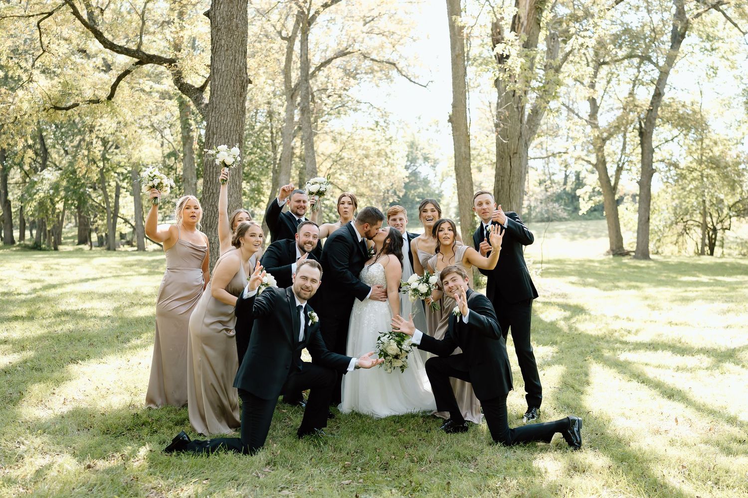 Wedding party poses playfully in a sunlit park setting with trees creating a natural backdrop.