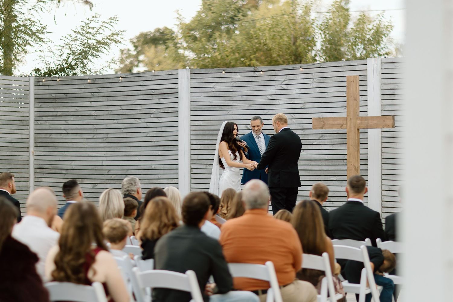 Wedding guests sit in white chairs during an outdoor ceremony with white fence panels in the background.