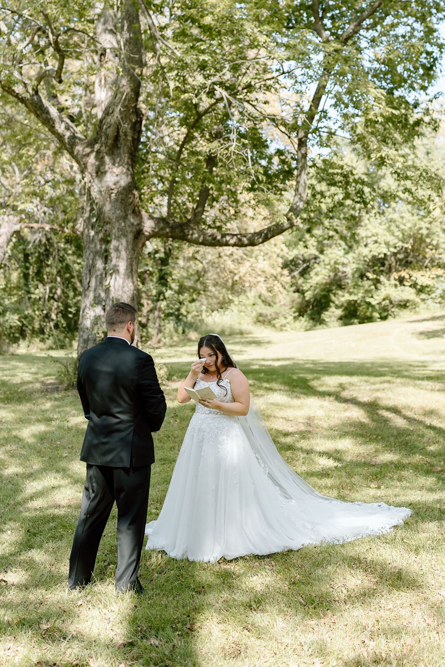 A couple exchanges vows during an intimate outdoor wedding ceremony beneath a large tree on a sunny summer day.