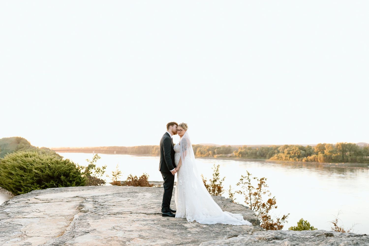 A couple in wedding attire stands at the edge of a lake during sunset with fall foliage in the background.