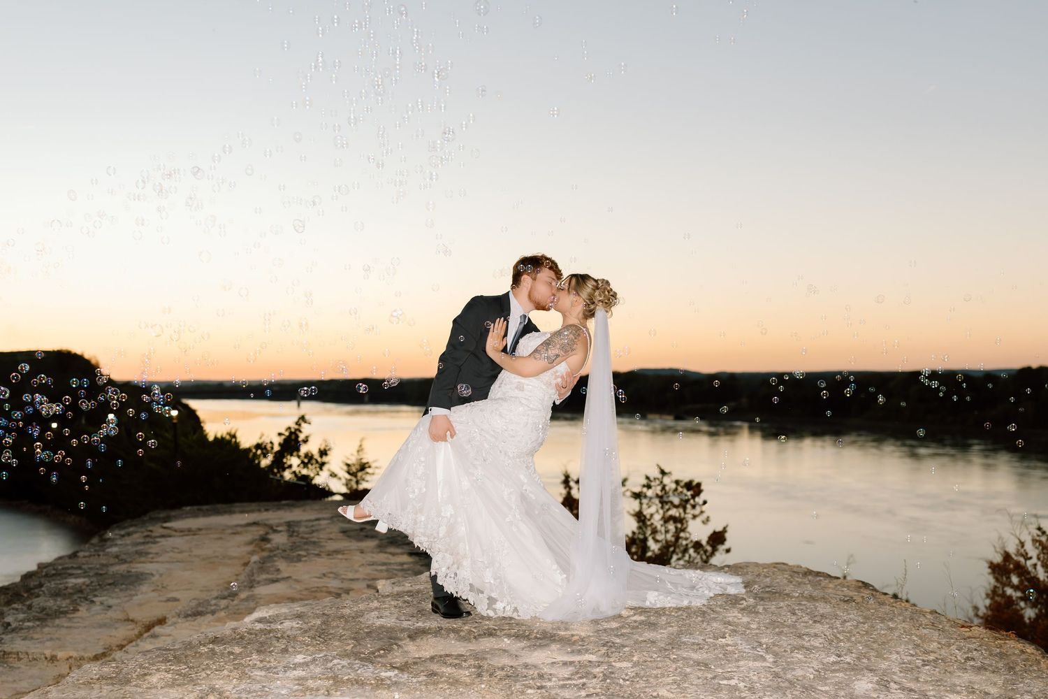 A couple in wedding attire shares a romantic moment at sunset overlooking a scenic lake.