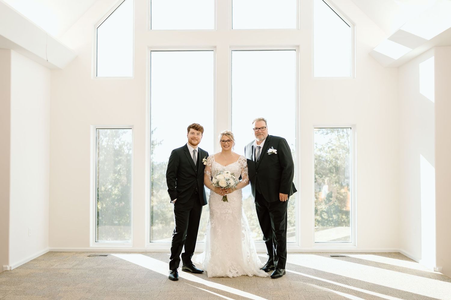 A wedding ceremony setup with five people standing in a bright white room with large windows and natural light.