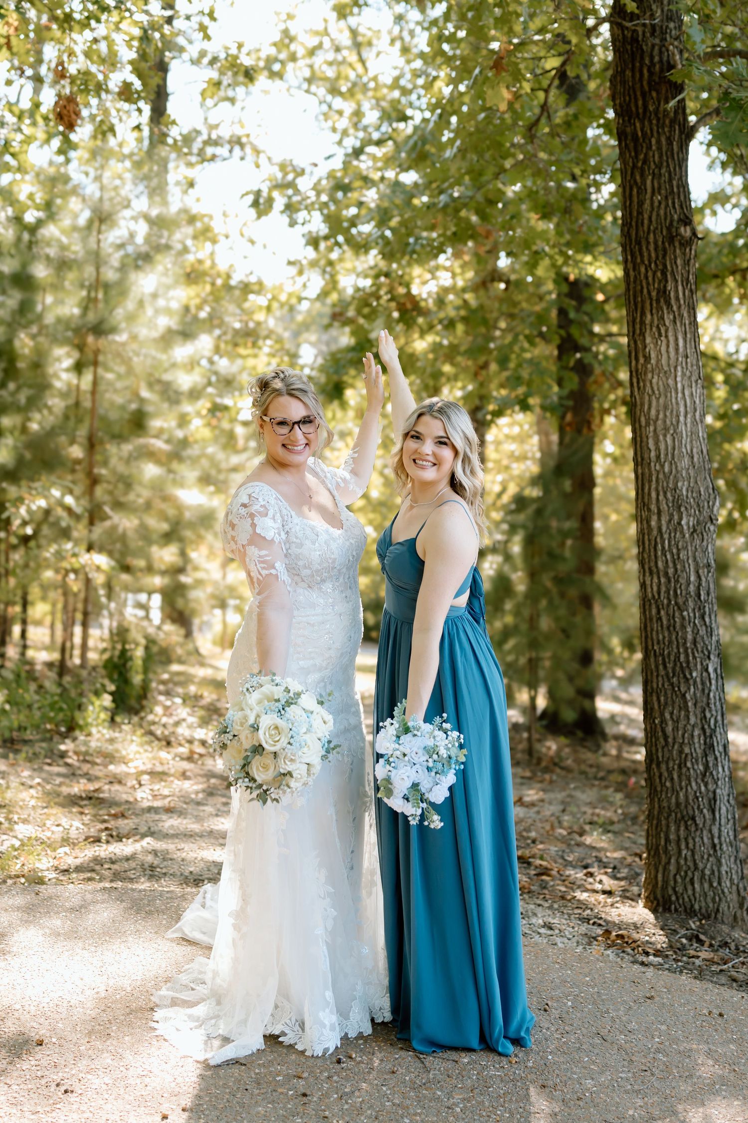 Two people in formal attire pose together on a wooded path, holding blue and white floral bouquets.
