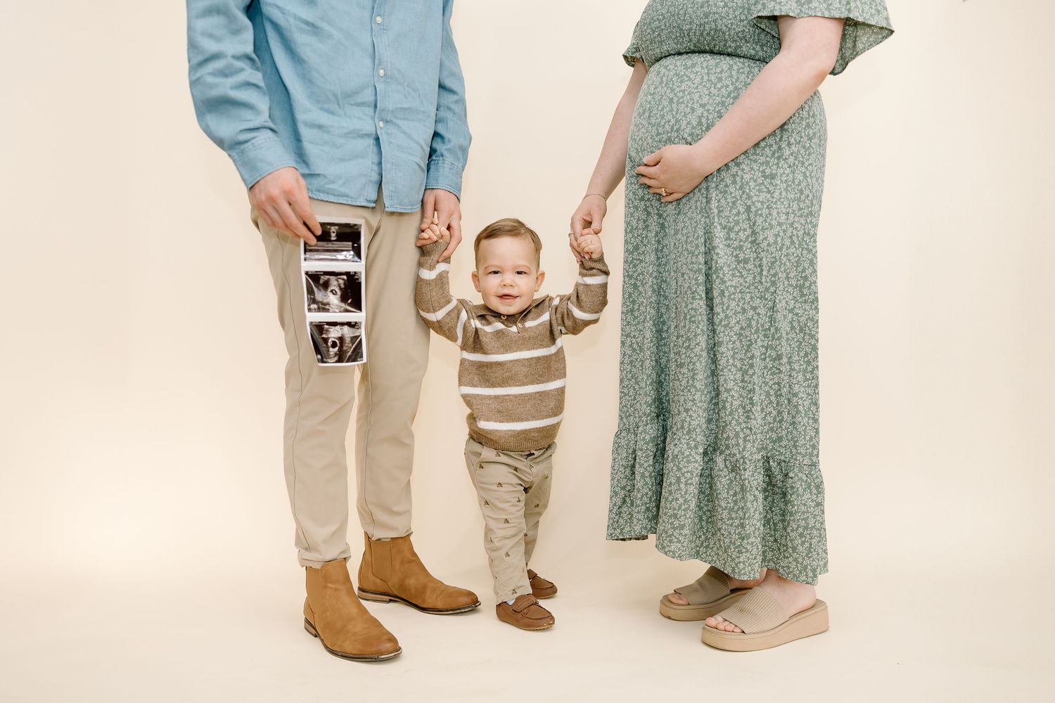 A family poses together against a beige backdrop while holding an ultrasound photo, announcing a new addition.