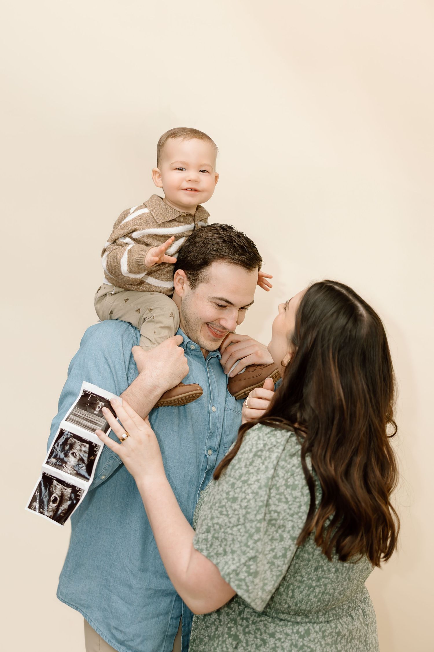 A couple shares joyful moments with a baby while holding ultrasound photos against a neutral background.