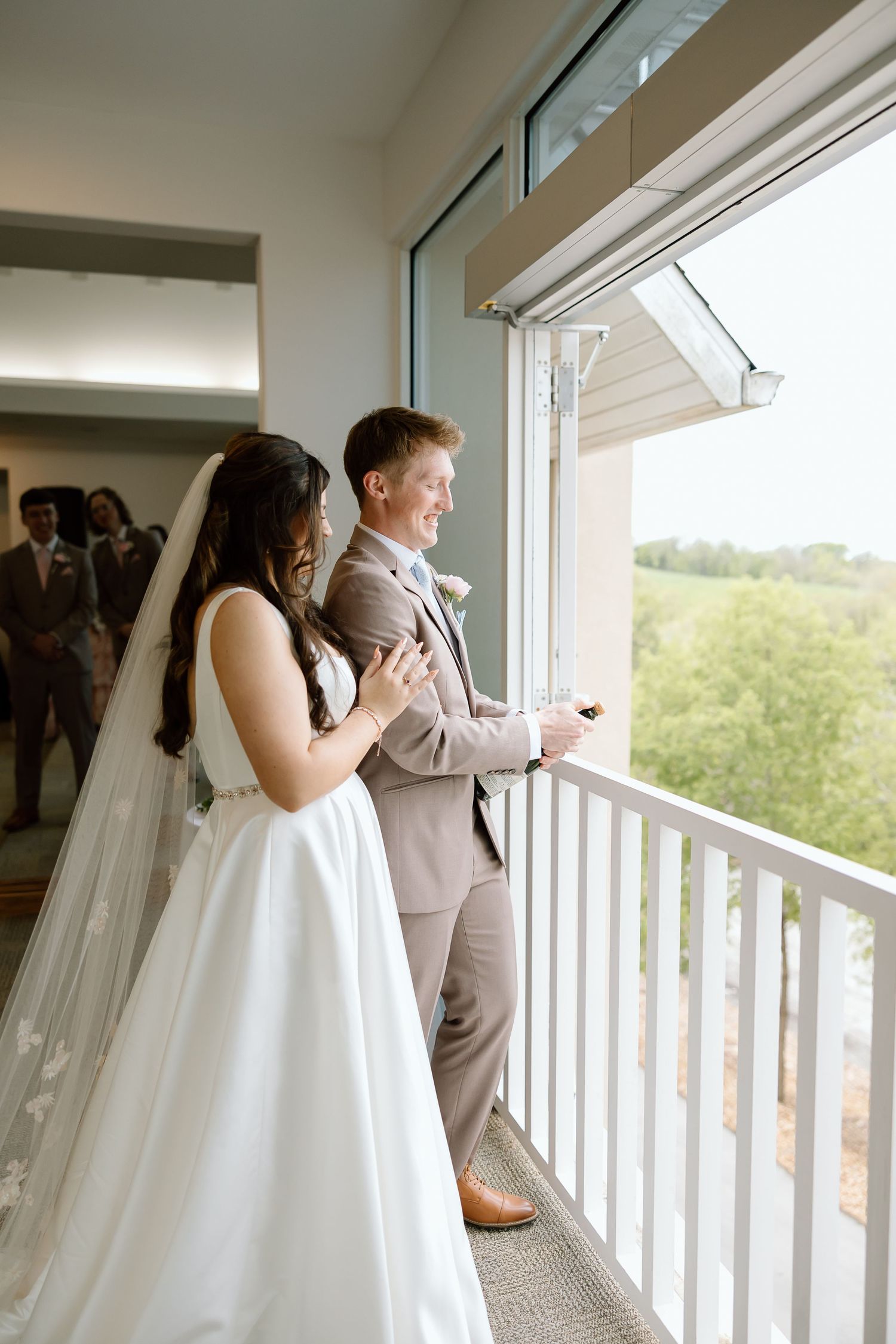 A bride in white gown and groom in tan suit share a moment together on a balcony overlooking green scenery.