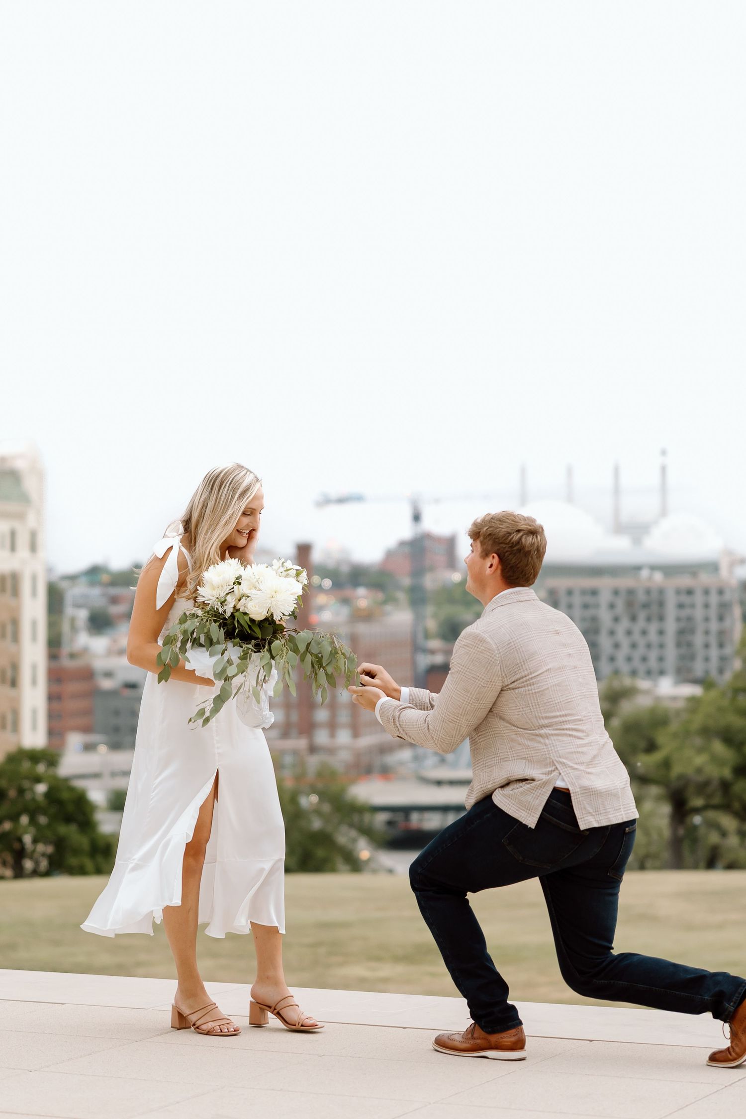 A romantic proposal moment captured on a hilltop overlooking a cityscape, with white flowers and formal attire.