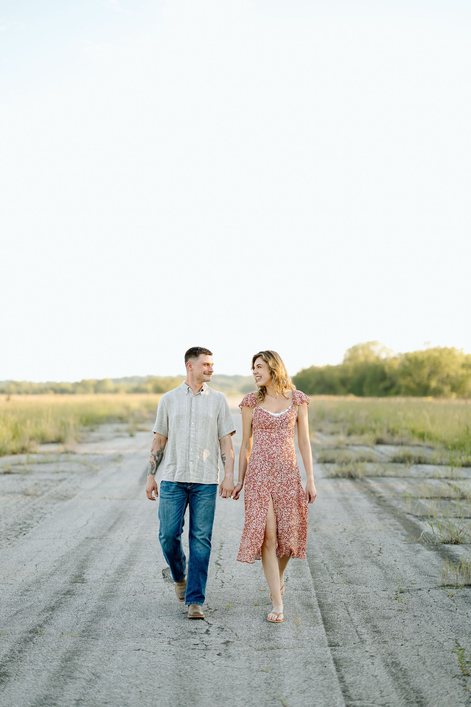 Couple walking hand in hand on a dirt path through a rural field at sunset.