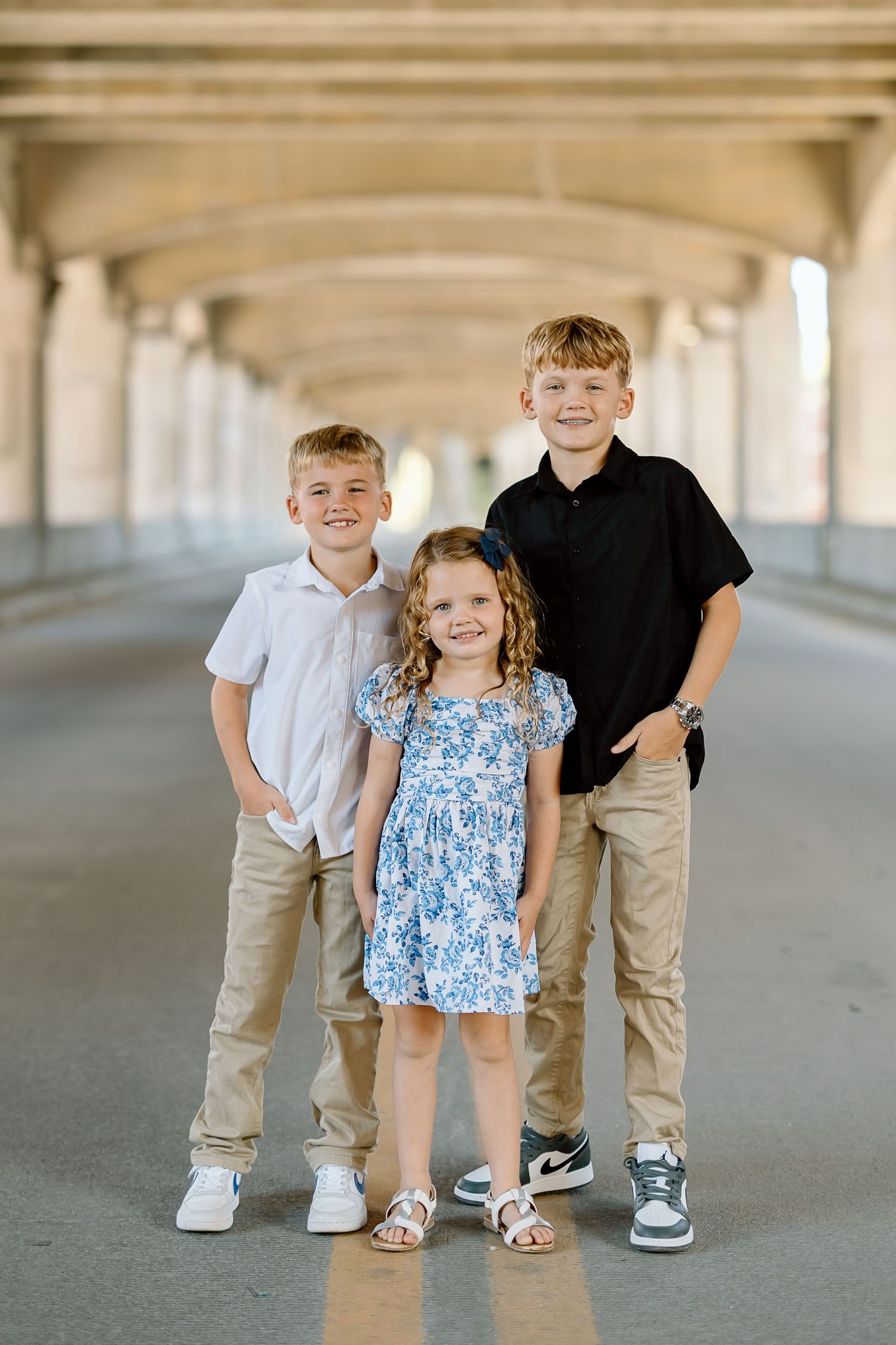 Three children in casual attire stand together under a covered walkway with concrete columns.