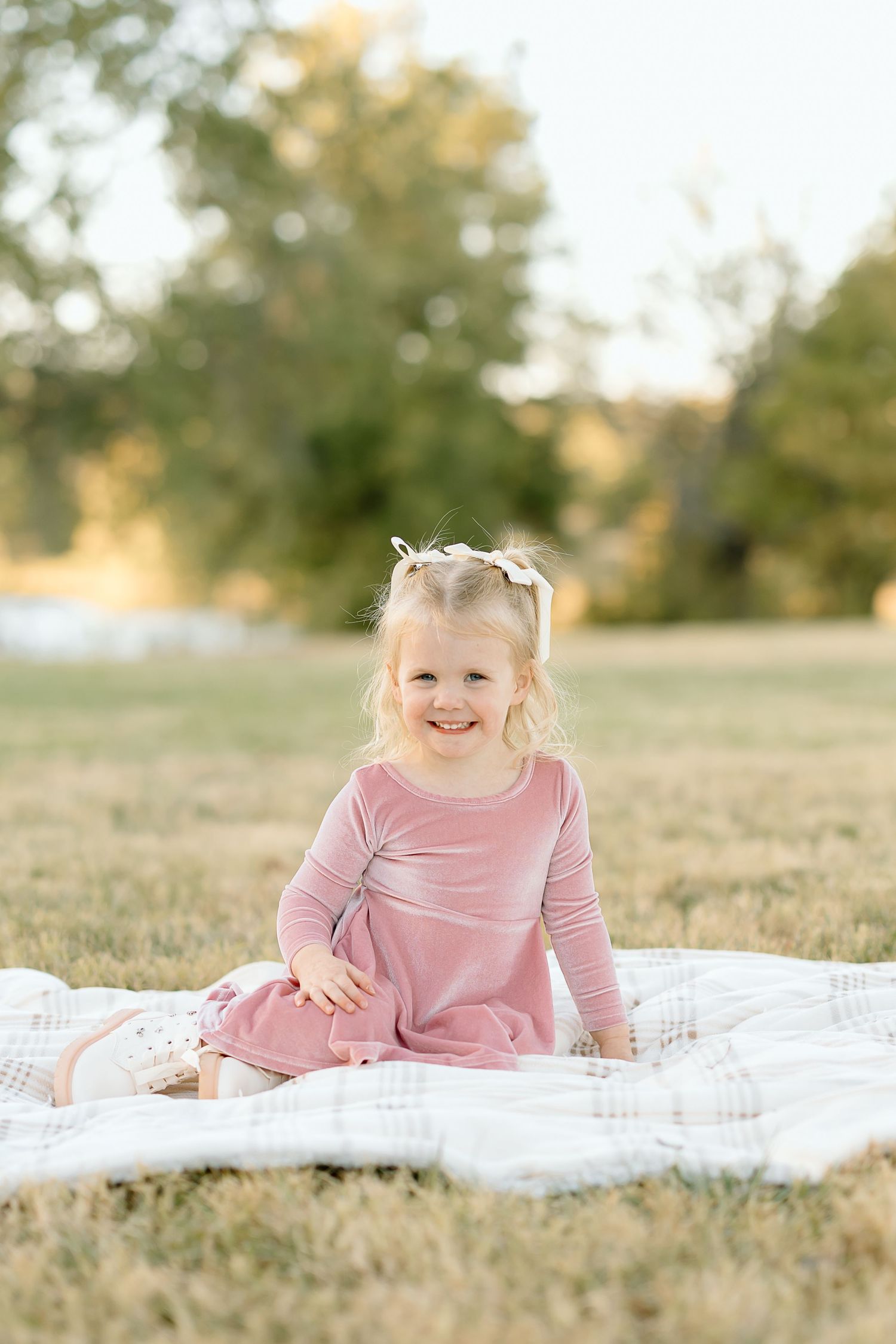 Three connected photos show a child in a pink dress lying on a white blanket in a grassy field at sunset.