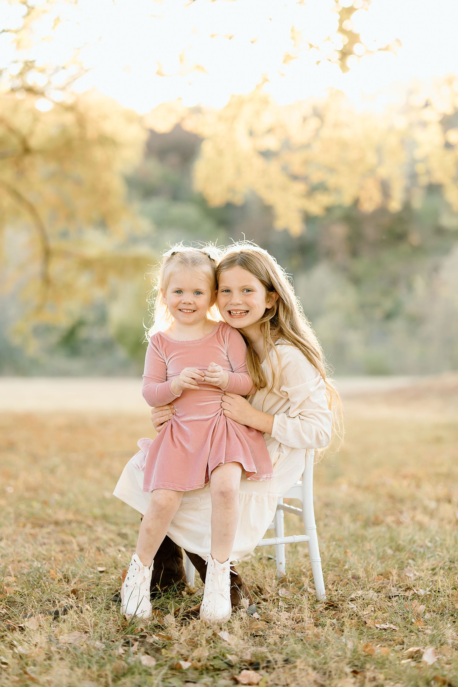 Two children in pink and white outfits sit together on a white chair in an autumn outdoor setting.