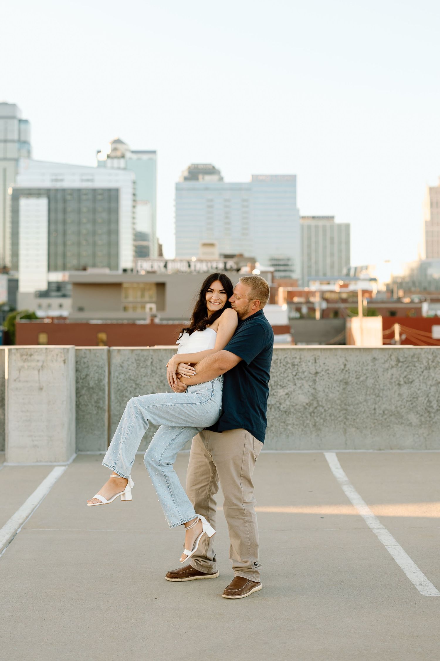 A couple shares a romantic moment in an urban rooftop parking lot with city buildings in the background at sunset.