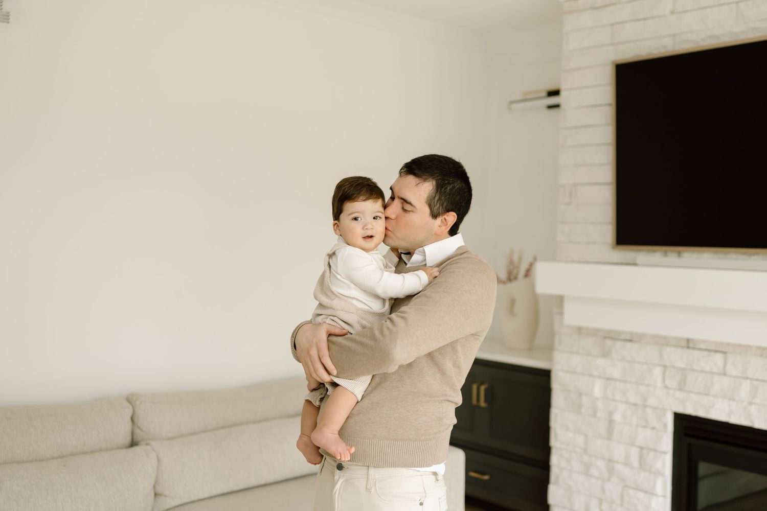 A tender family moment captured in black and white against a white brick wall with a mounted TV screen.