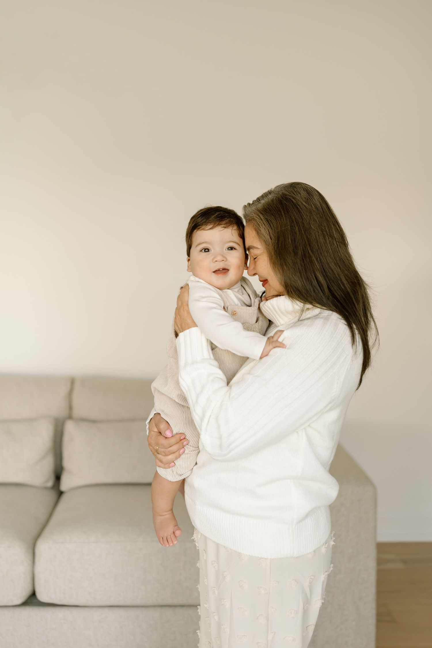 A tender moment between parent and baby in white clothing standing by a grey couch in a minimalist room.