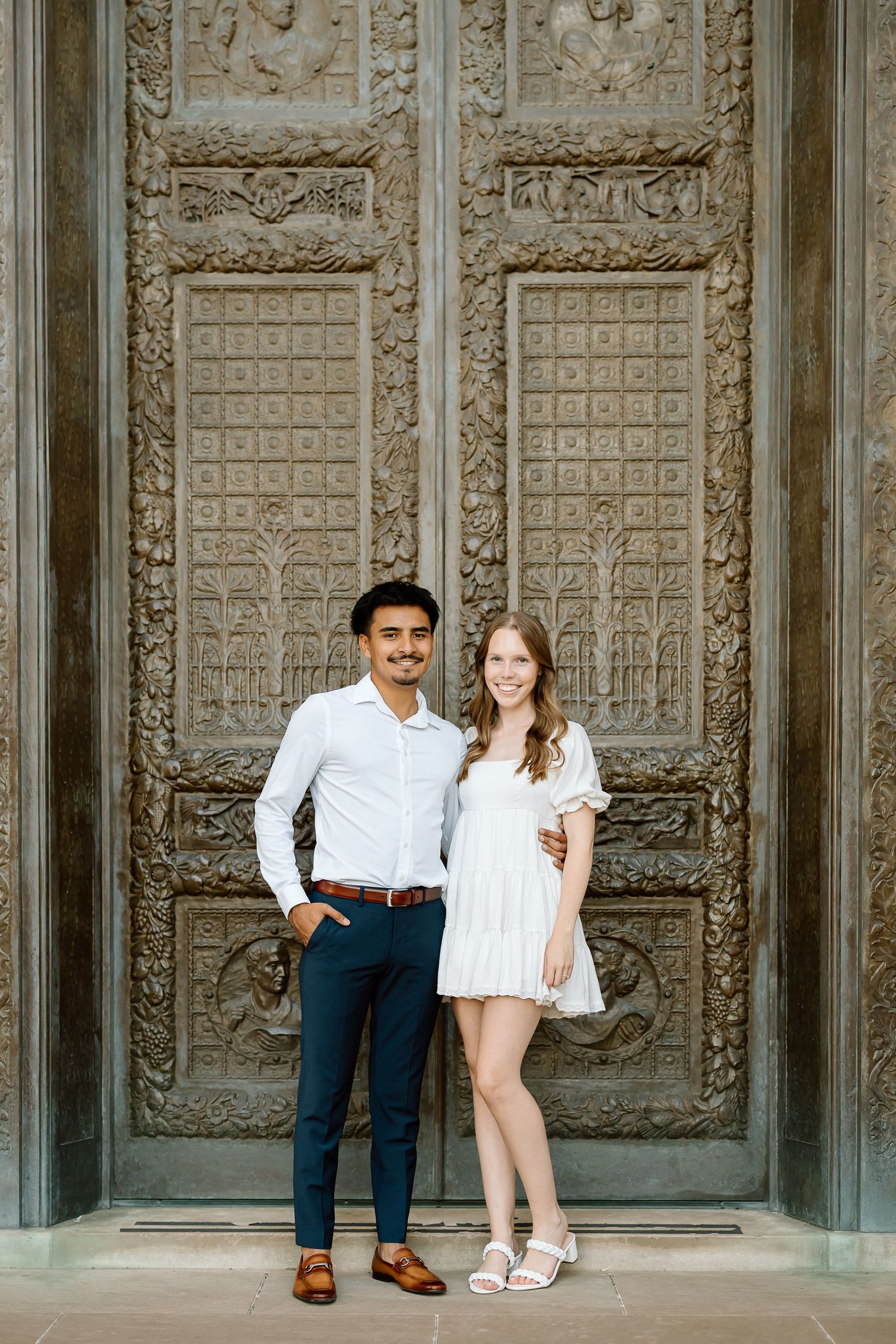A couple poses casually against an ornate carved stone wall with decorative architectural details and panels.
