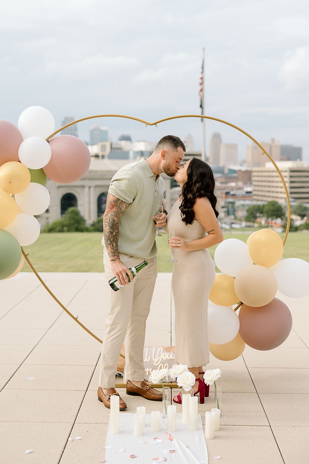 Romantic rooftop proposal setup with heart-shaped balloon arch, candles and city skyline backdrop.