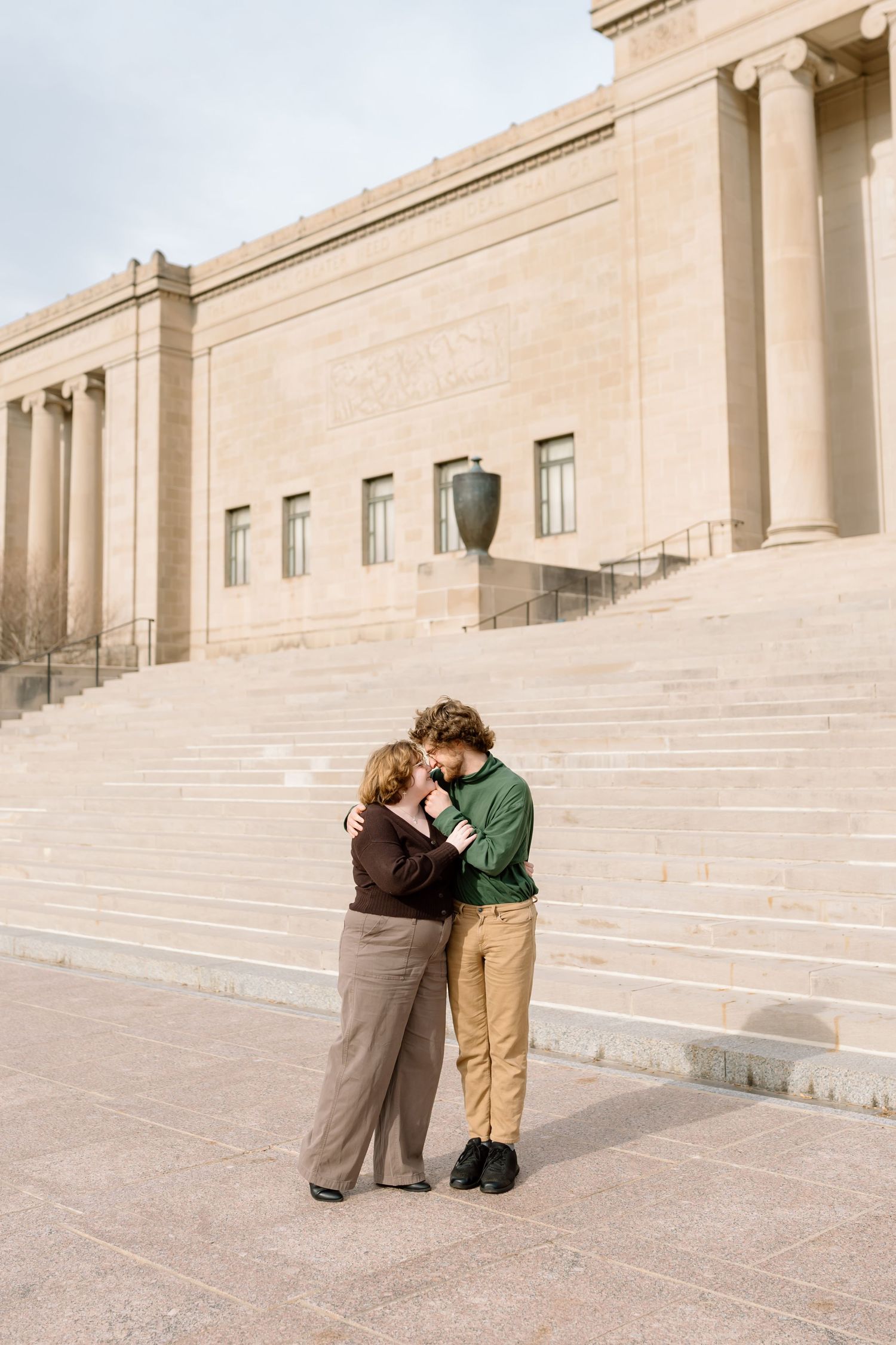 A couple embraces on the grand stone steps of a neoclassical building with columns and beige facade.