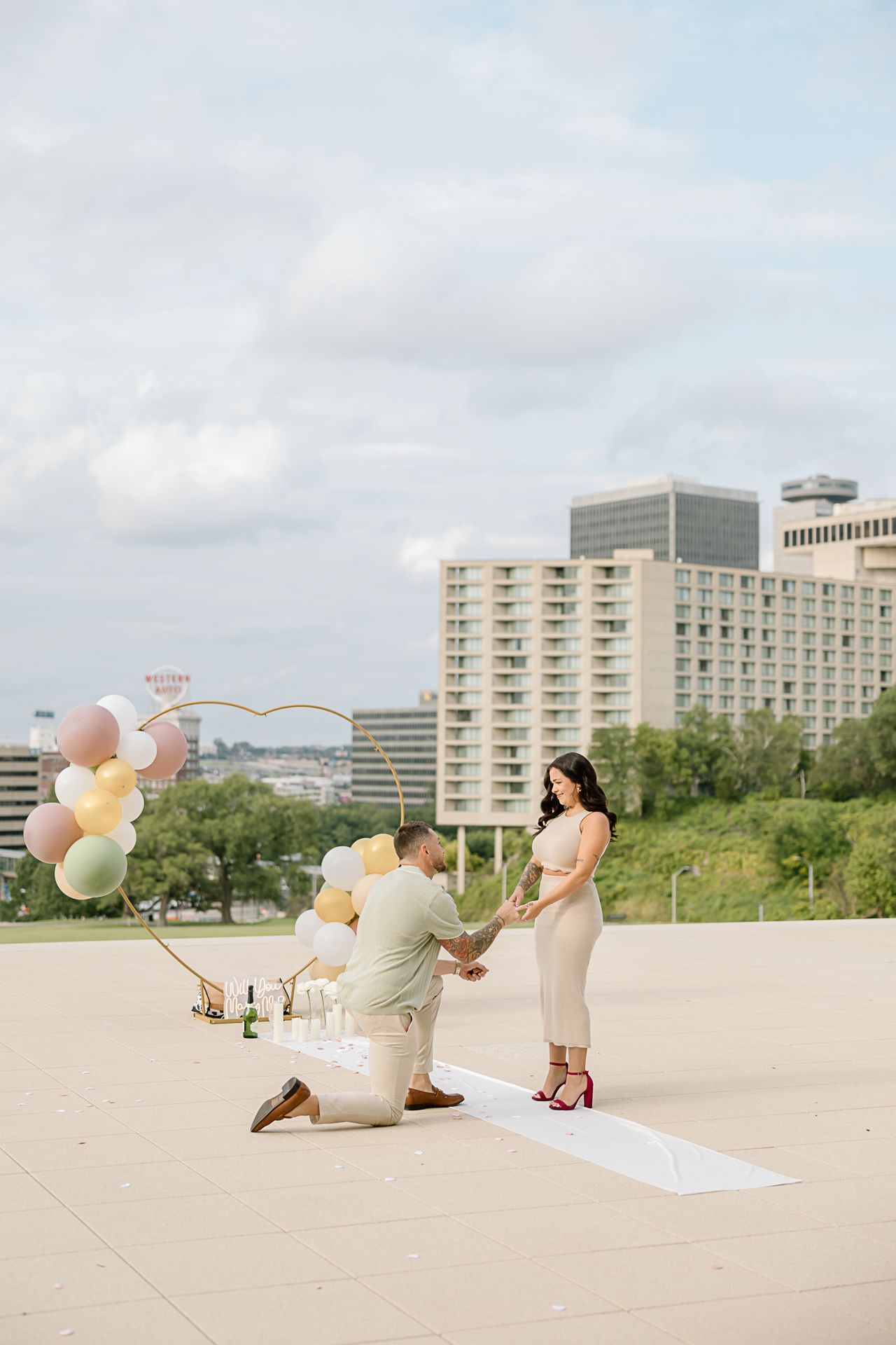 Romantic rooftop proposal scene with balloons and decorations against an urban skyline.