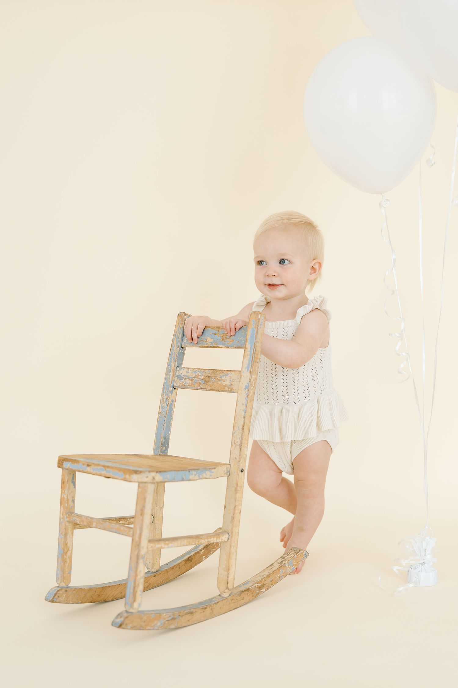 A toddler holds onto wooden rocking chair with white balloon floating nearby against cream backdrop.