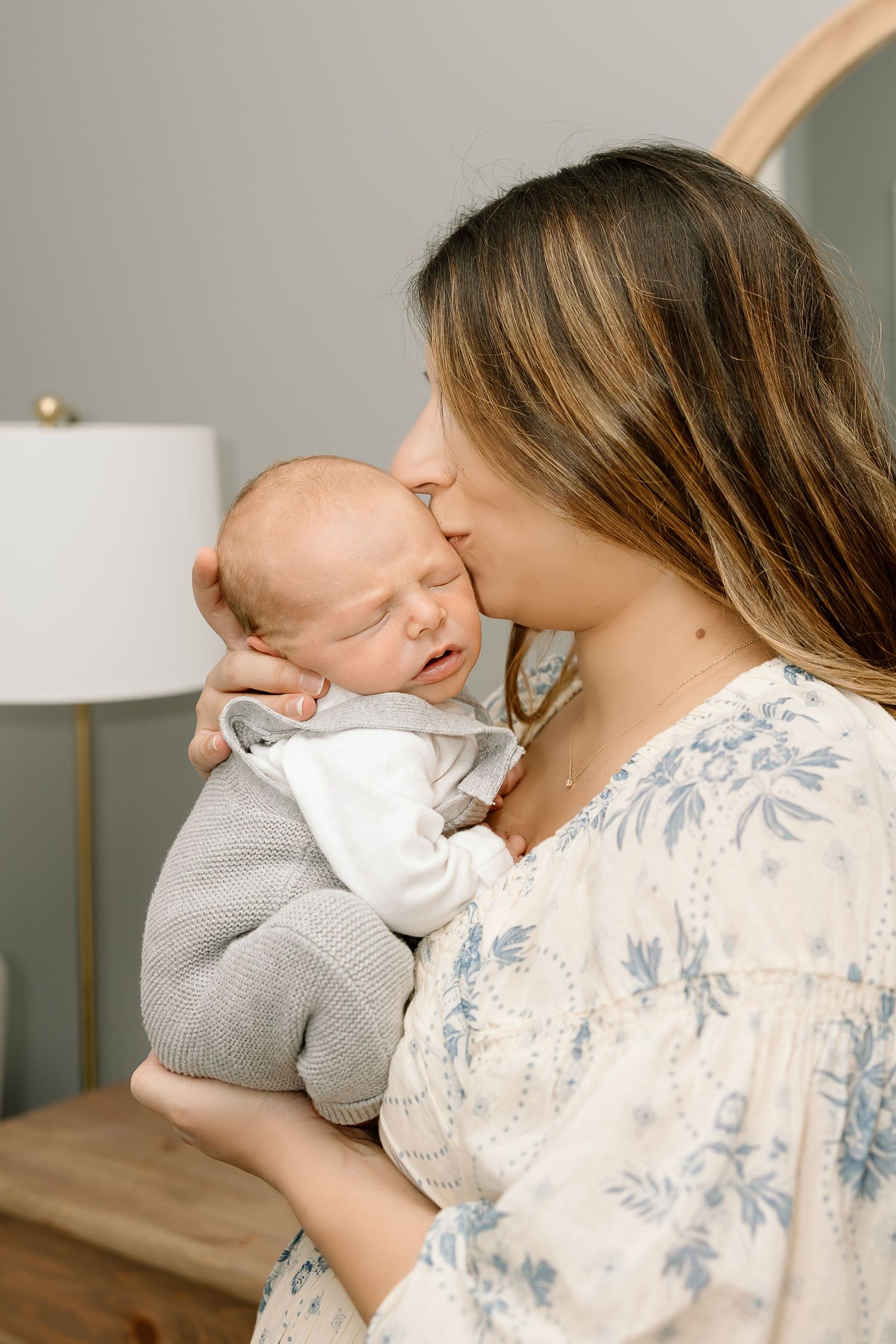 Parent holds sleeping baby wearing gray outfit while wearing floral print top in soft natural lighting.