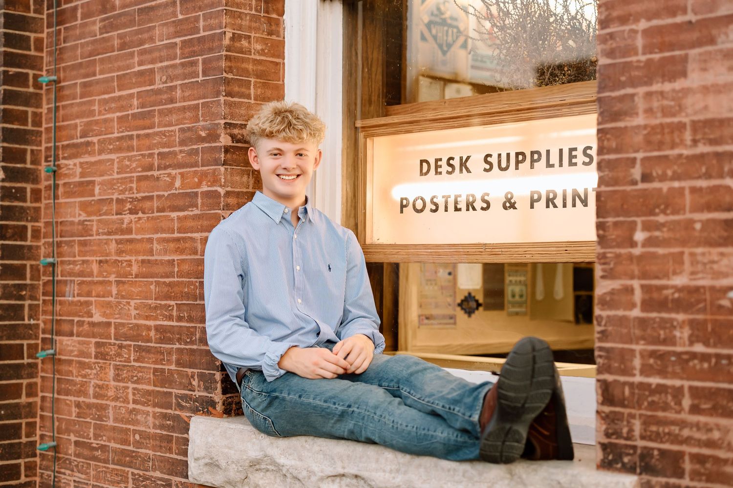 Someone sits casually on a brick windowsill outside a desk supplies and posters store in a light blue button-down shirt.