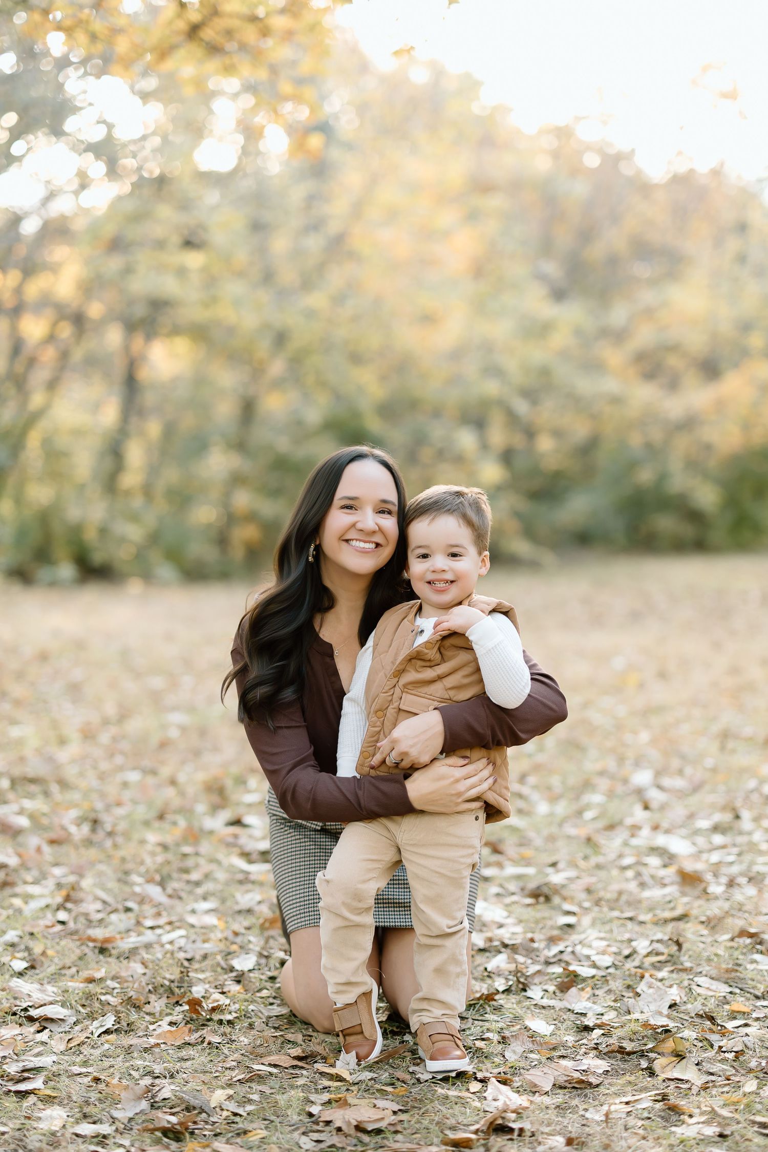 A mother and child share a joyful moment together outdoors during autumn with fallen leaves on the ground.