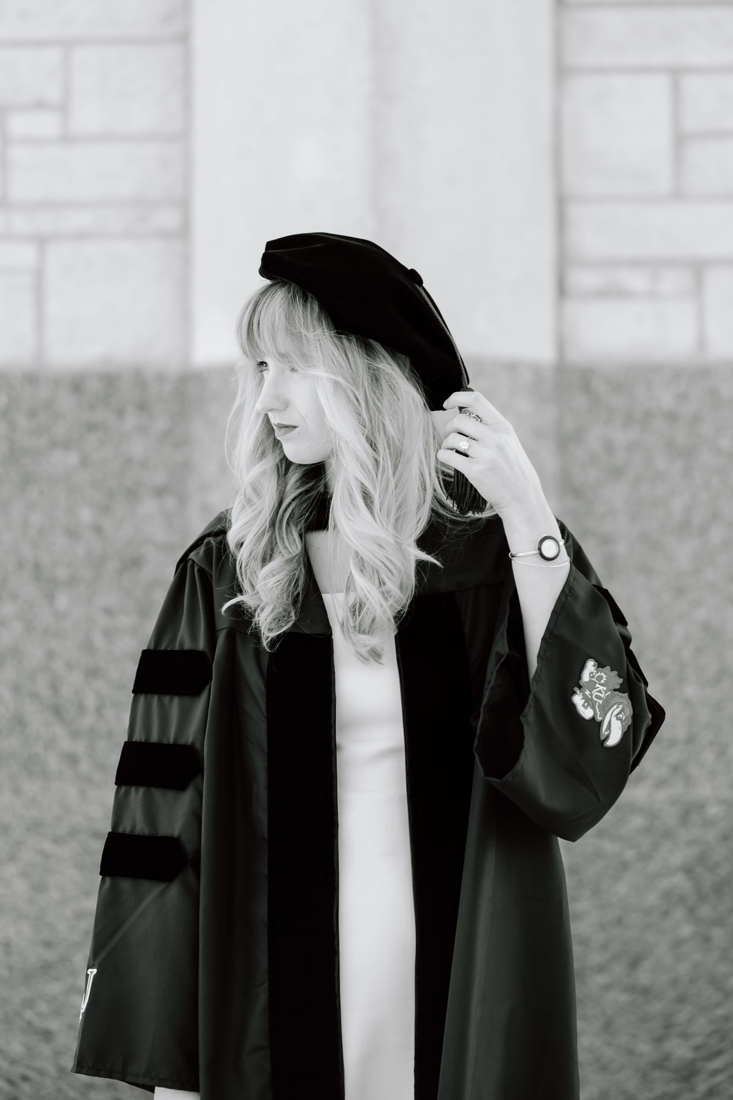 A black and white photo series showing someone wearing a black graduation gown and cap against a textured wall.