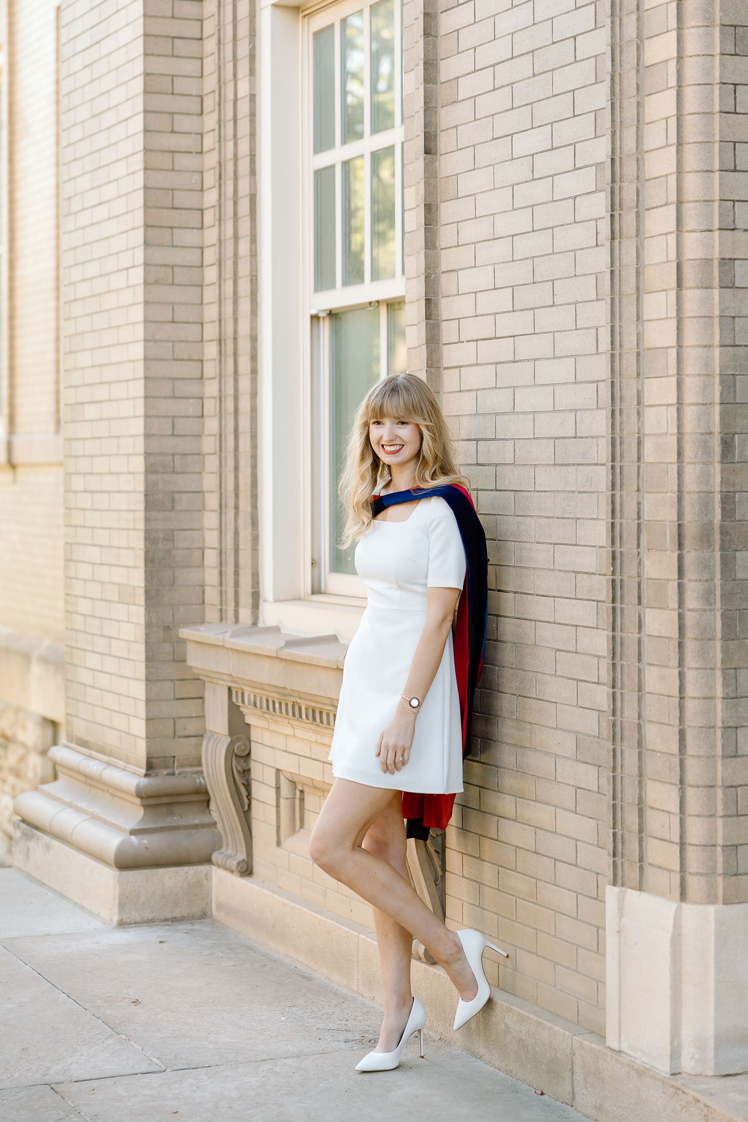A person in a white dress and sneakers poses playfully against a light brick building with columns and windows.