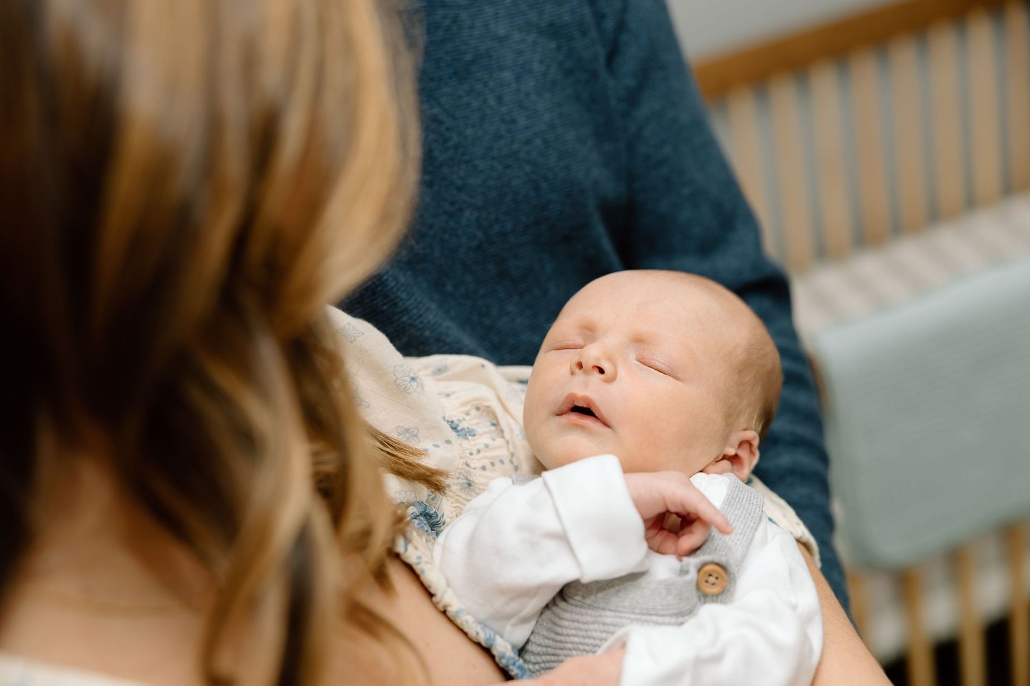 A baby yawns while being held in someone's arms wearing a white onesie.