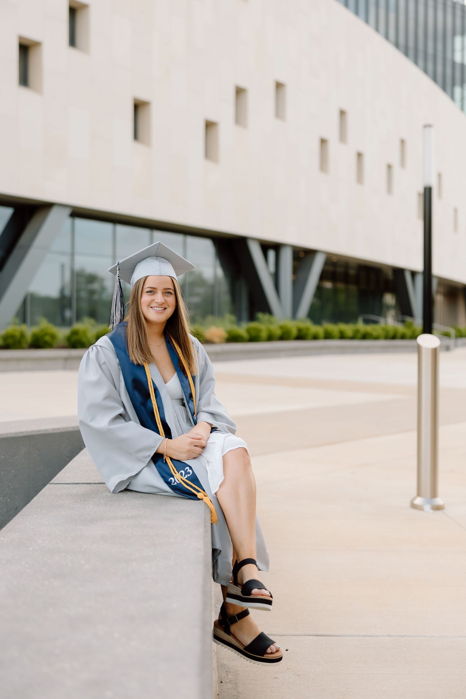 Graduate in cap and gown sits on concrete steps during graduation photoshoot at modern building.