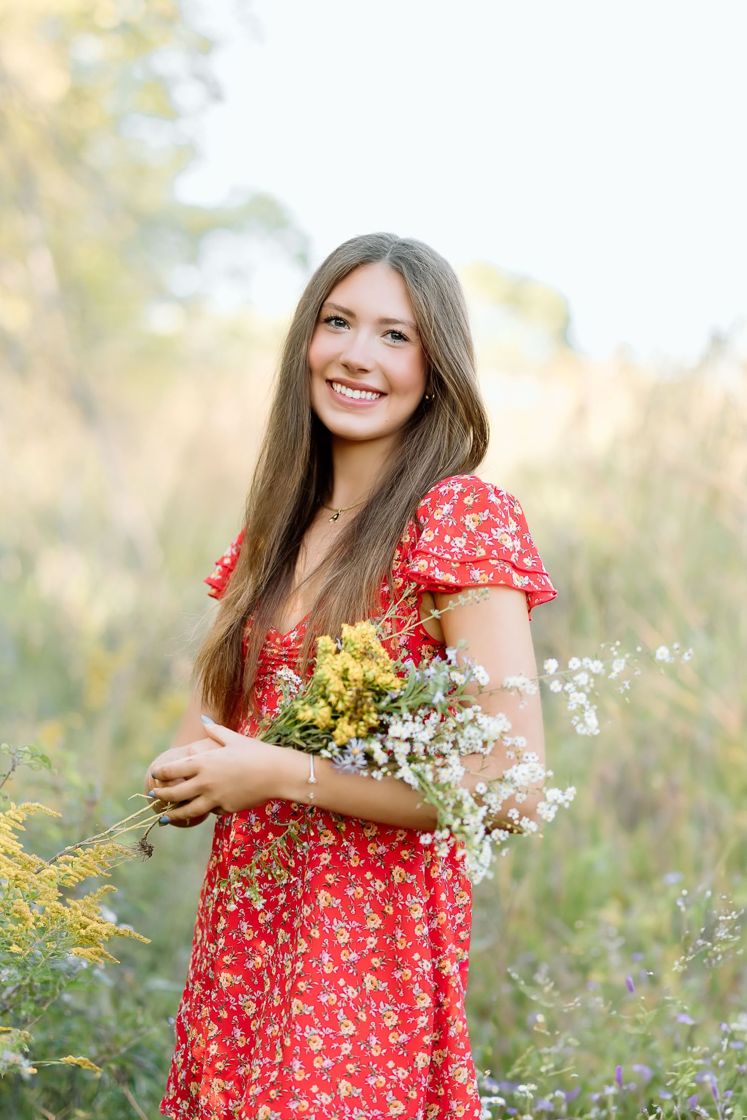 A woman in a red floral dress holds wildflowers while standing in a sunlit meadow during golden hour.