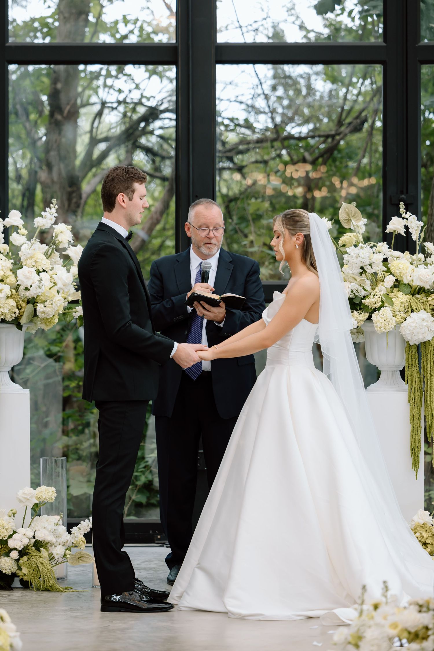Bride and groom exchange vows at elegant indoor wedding ceremony with white floral arrangements and large glass windows.