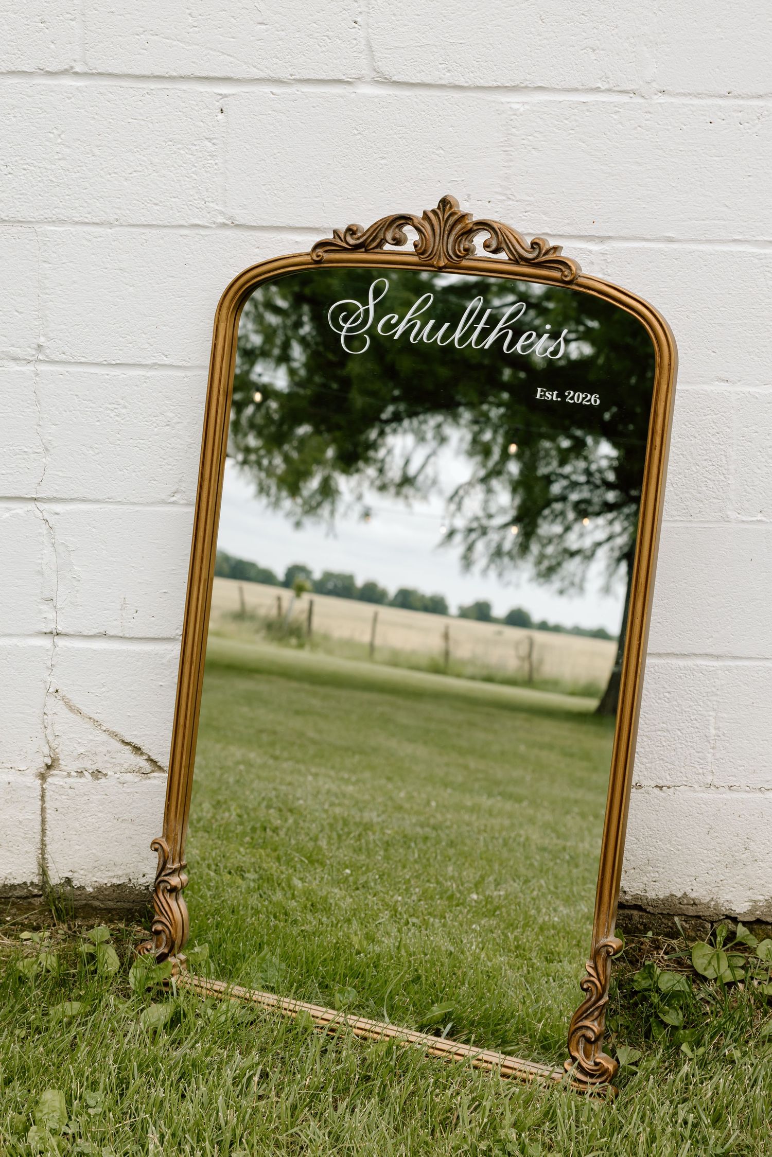 Gold ornate vintage mirror leaning against white brick wall displaying Schultheis Est. 2026 wedding signage.