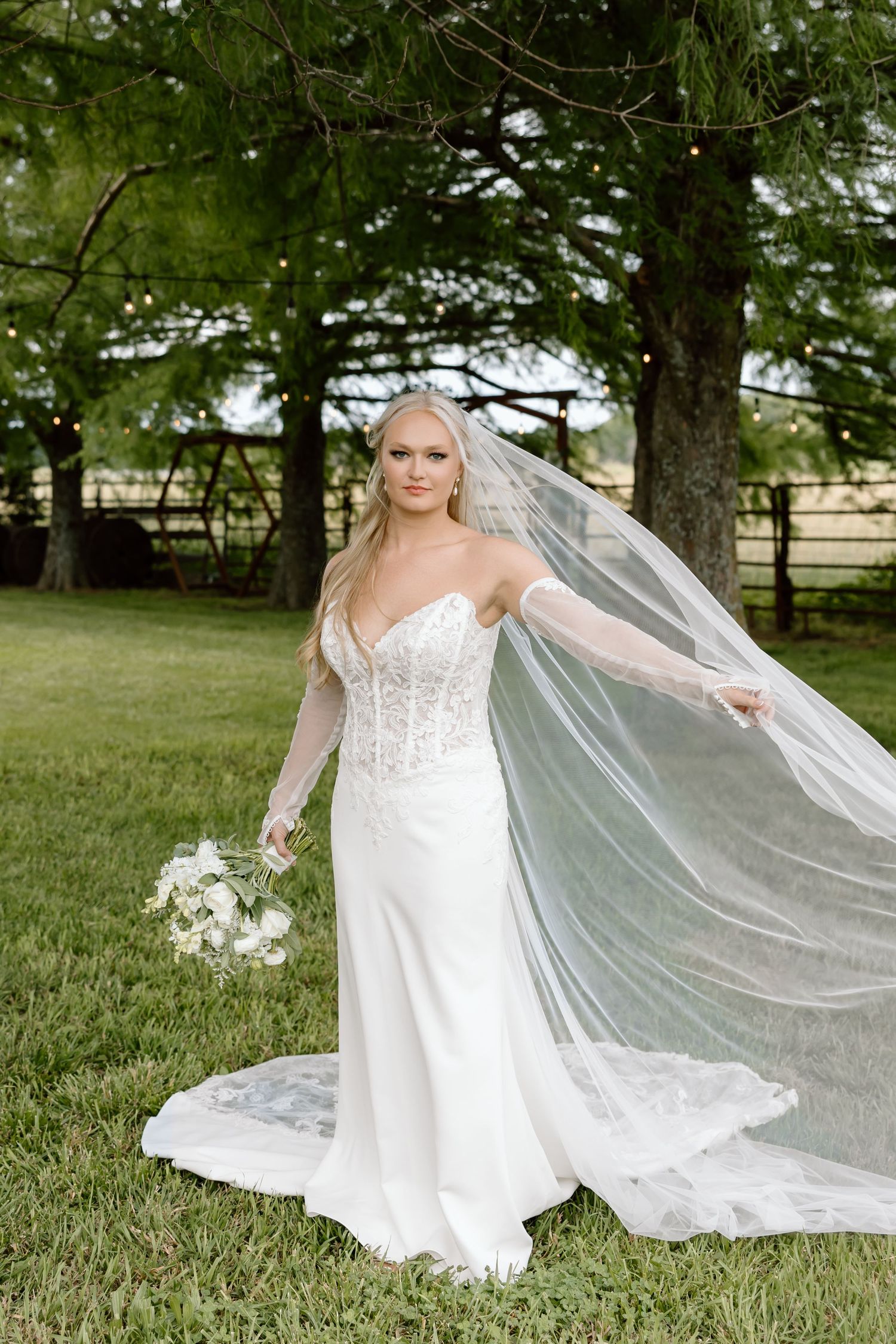 Bride in lace wedding gown with flowing veil holds bouquet outdoors on green lawn near trees.