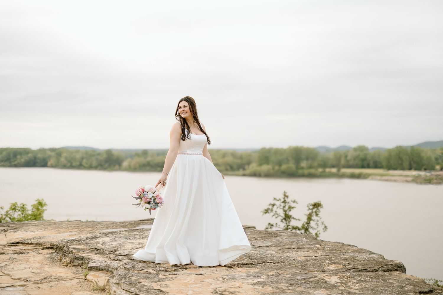 Bride in flowing white dress poses on rocky outcrop with lake and forest backdrop.