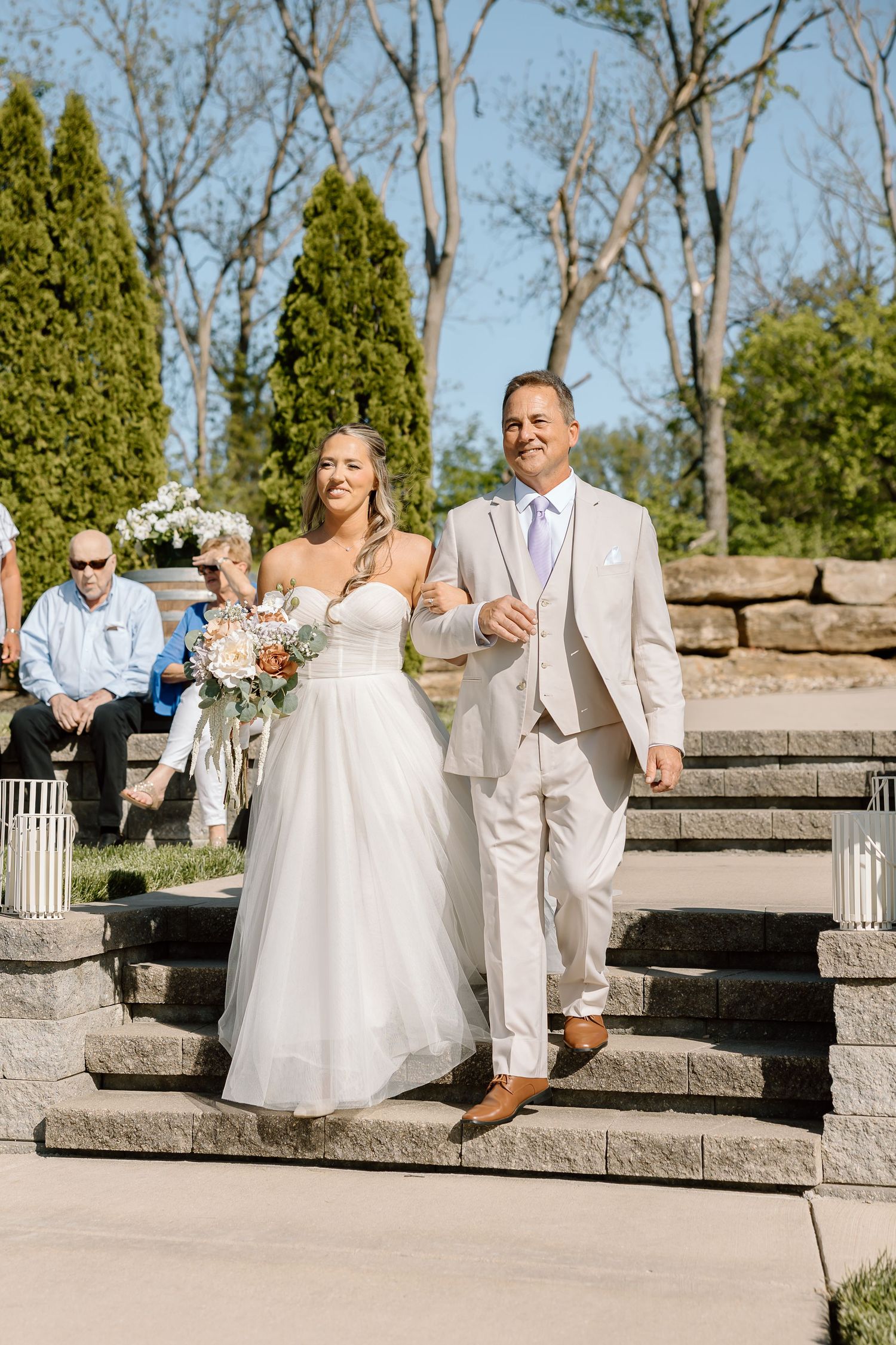Newlyweds descend stone steps after ceremony with bride in white gown and groom in light beige suit.