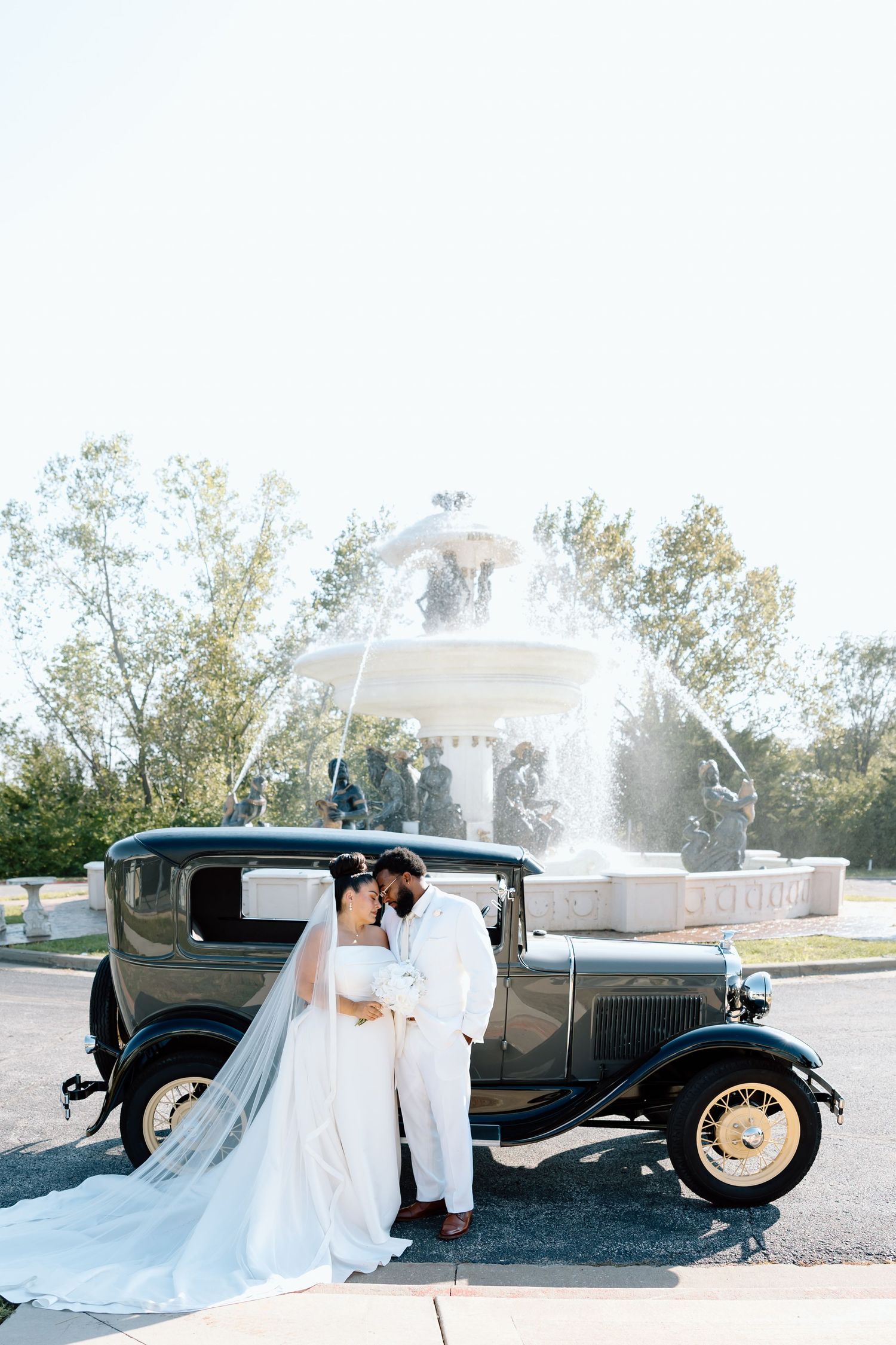 Bride and groom pose beside a vintage black car with a white fountain backdrop on their wedding day.