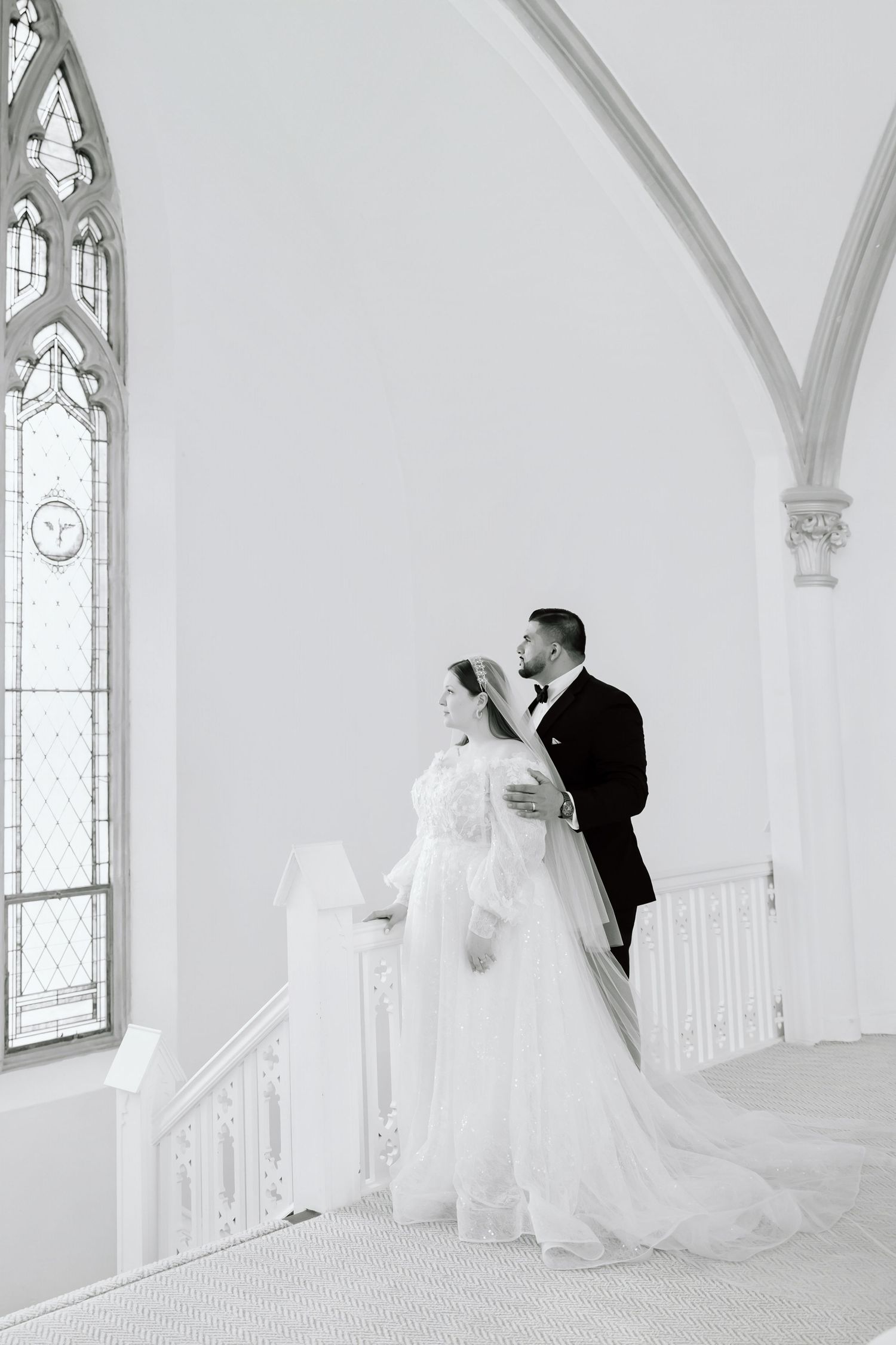 Black and white photo of bride and groom standing together on church staircase with gothic arched windows.