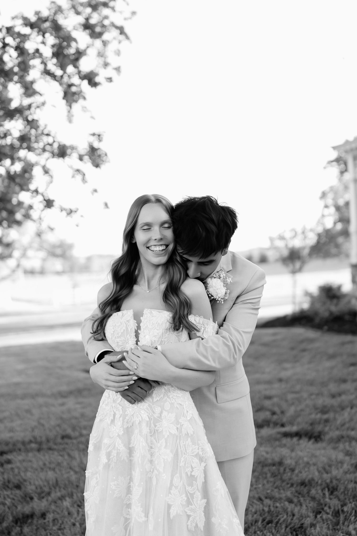 Black and white photo of a smiling bride in lace gown embraced by groom in tan suit outdoors.