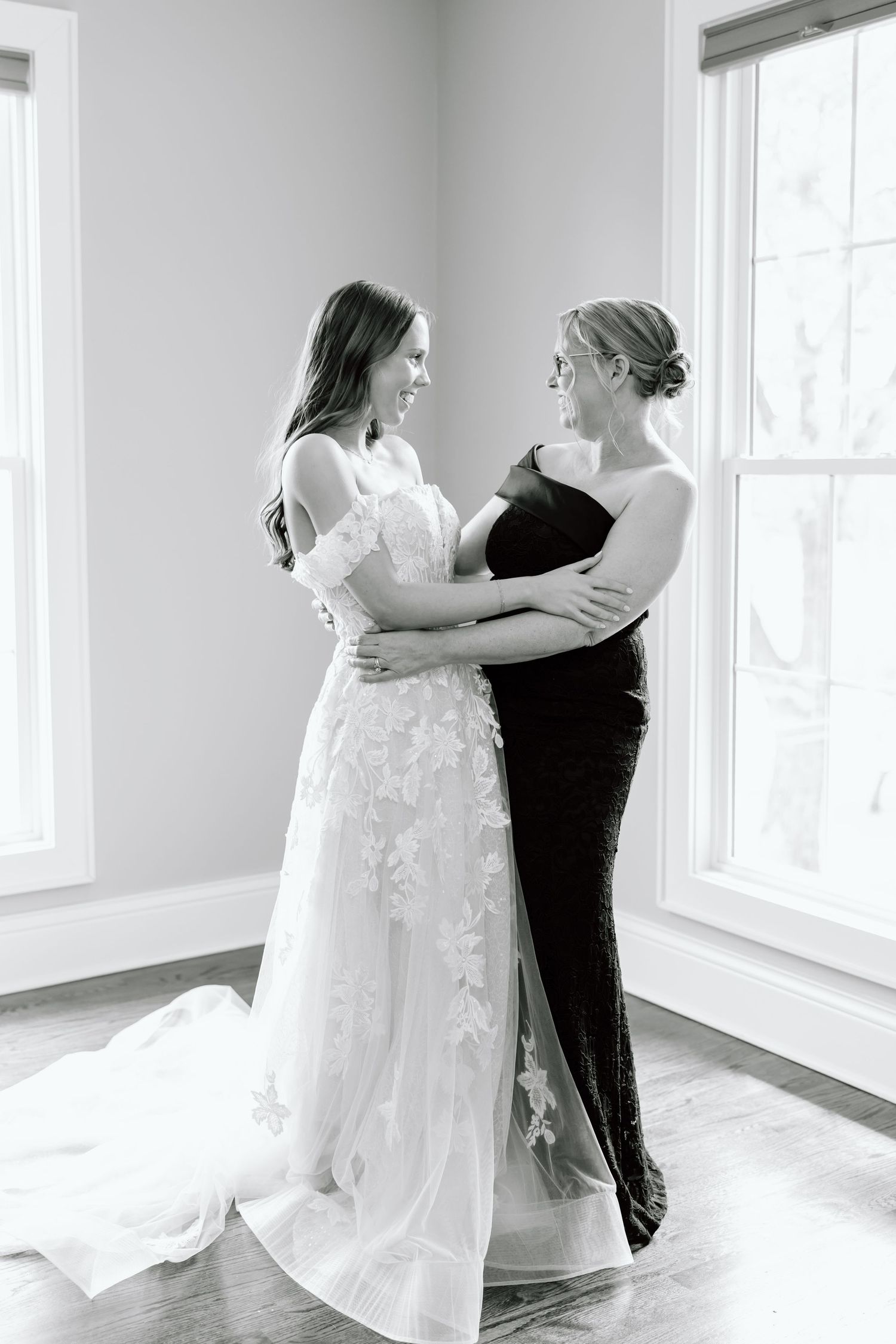Black and white photo of a bride in lace gown embracing a woman in dark dress, sharing a joyful moment by sunlit windows.