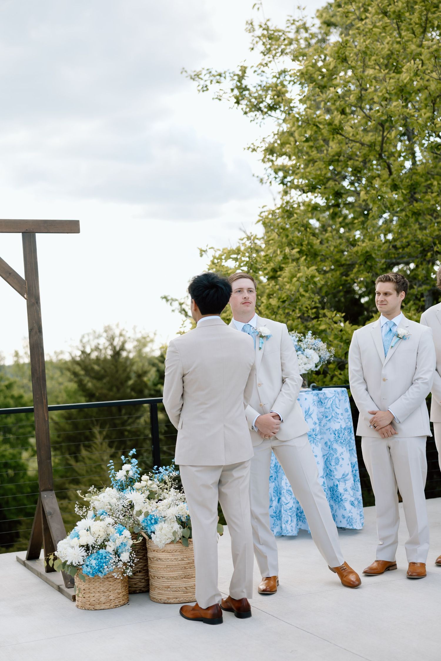 Groom and groomsmen in tan suits with blue ties stand at an outdoor wedding ceremony altar with white and blue floral arrangements.
