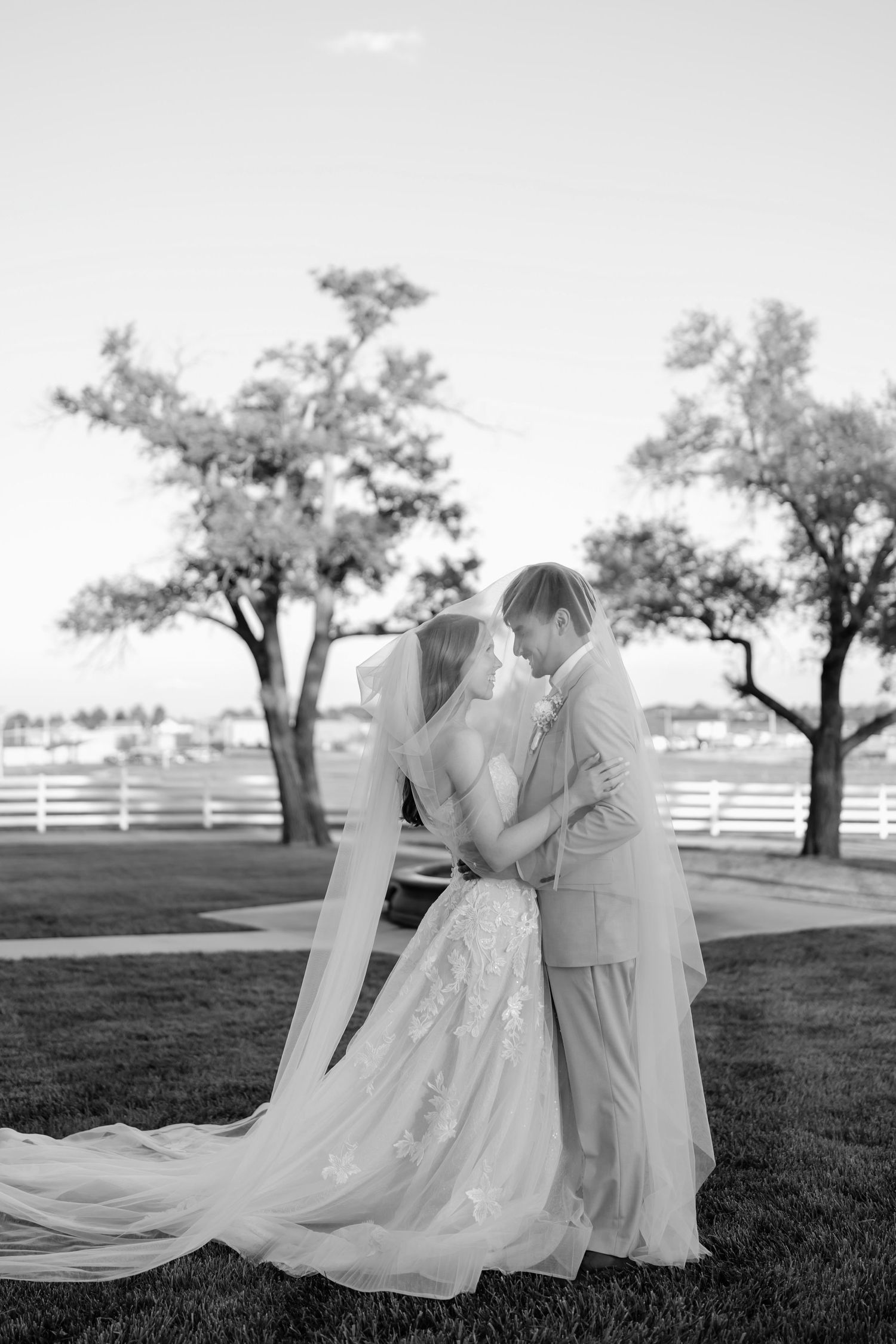 Black and white photo of bride and groom sharing an intimate moment under veil outdoors near white fence and trees.