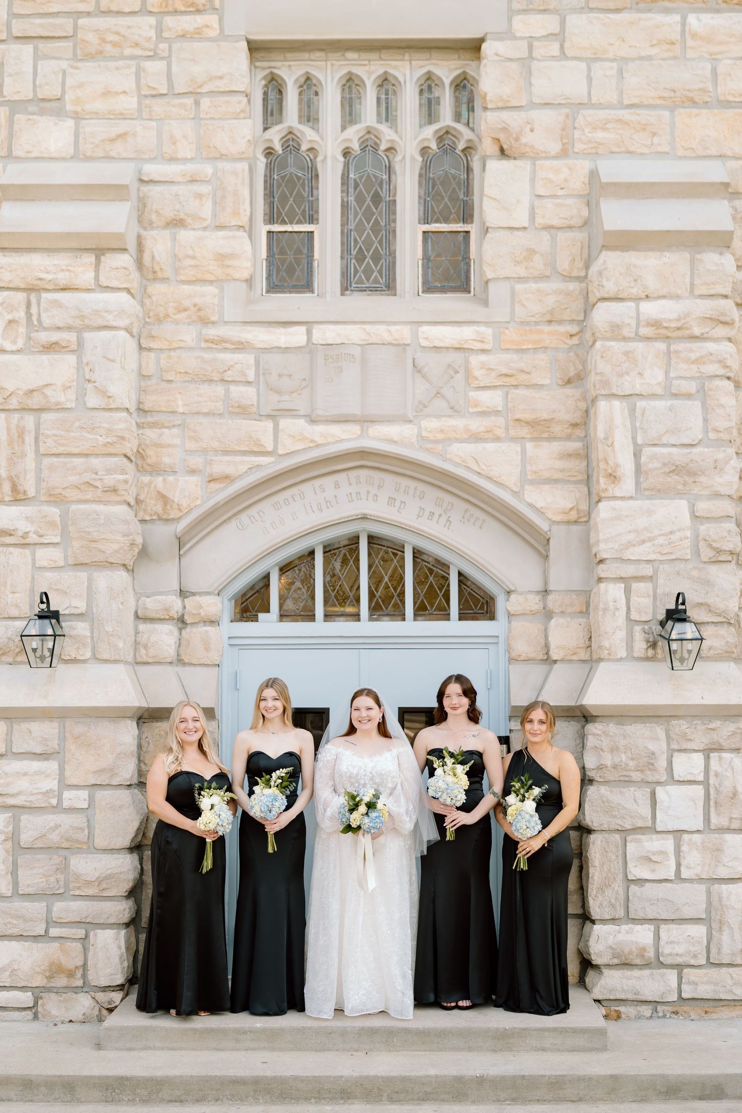 Bride in white gown with four bridesmaids in black dresses holding white bouquets outside a stone church.