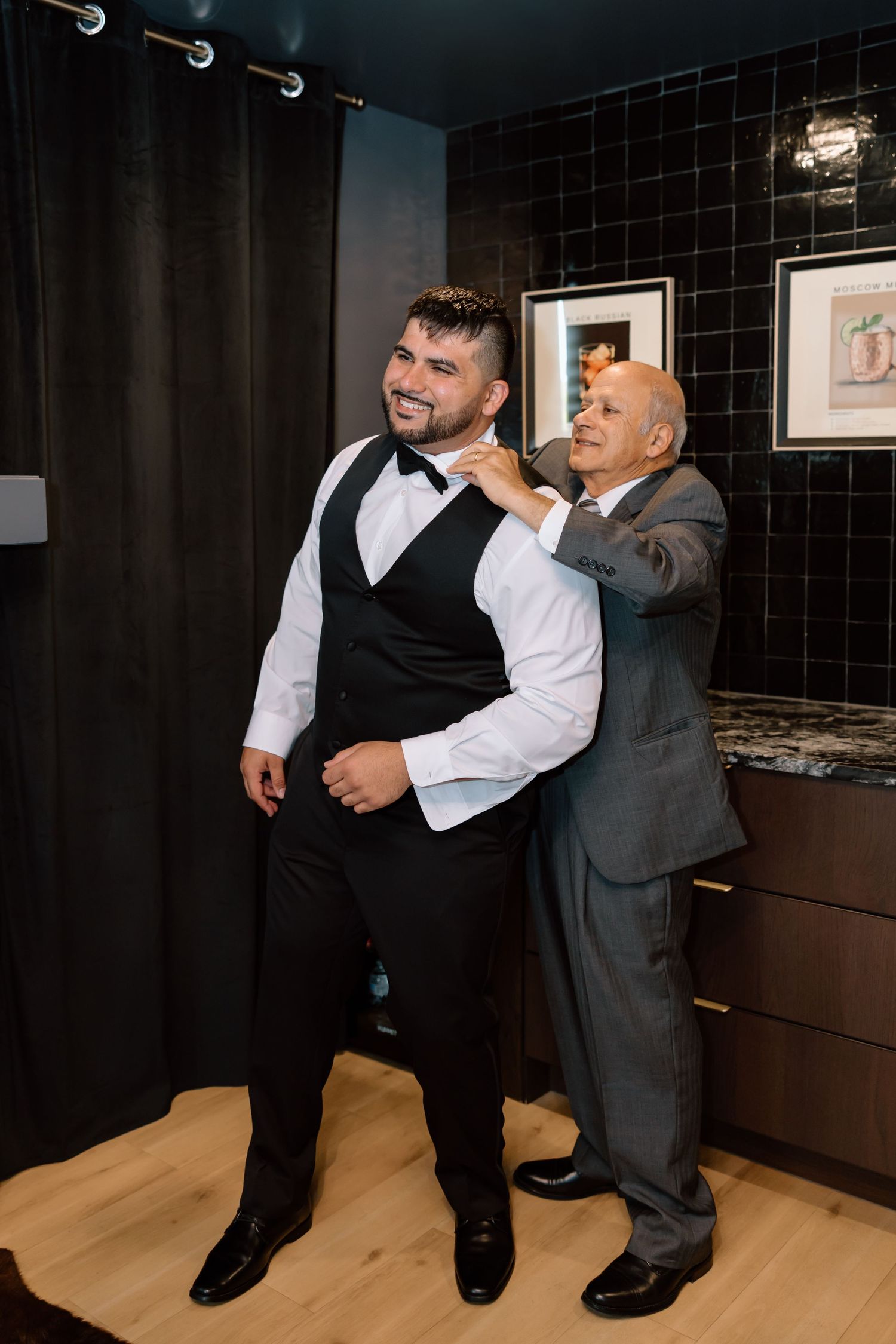 A smiling groom in a black vest and bow tie gets help adjusting his collar from an older man in a gray suit.