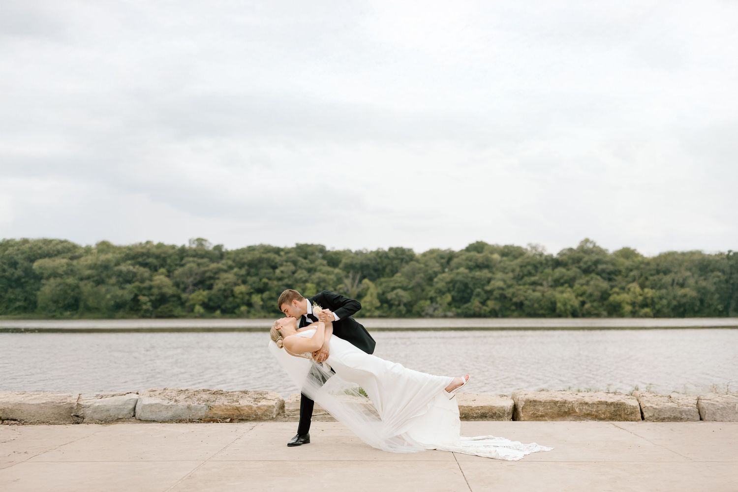 Two people in wedding attire running playfully along a scenic beach path with trees in the background.