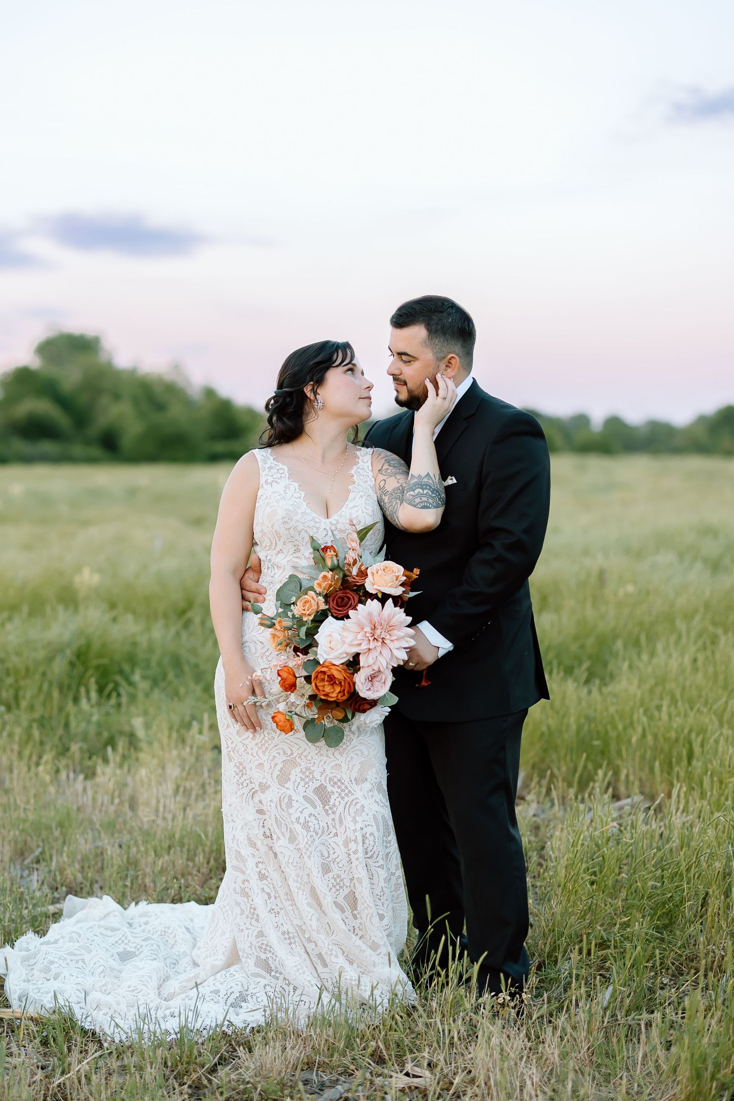 A bride and groom share romantic moments in a sunlit meadow with peach and coral florals in her bouquet.